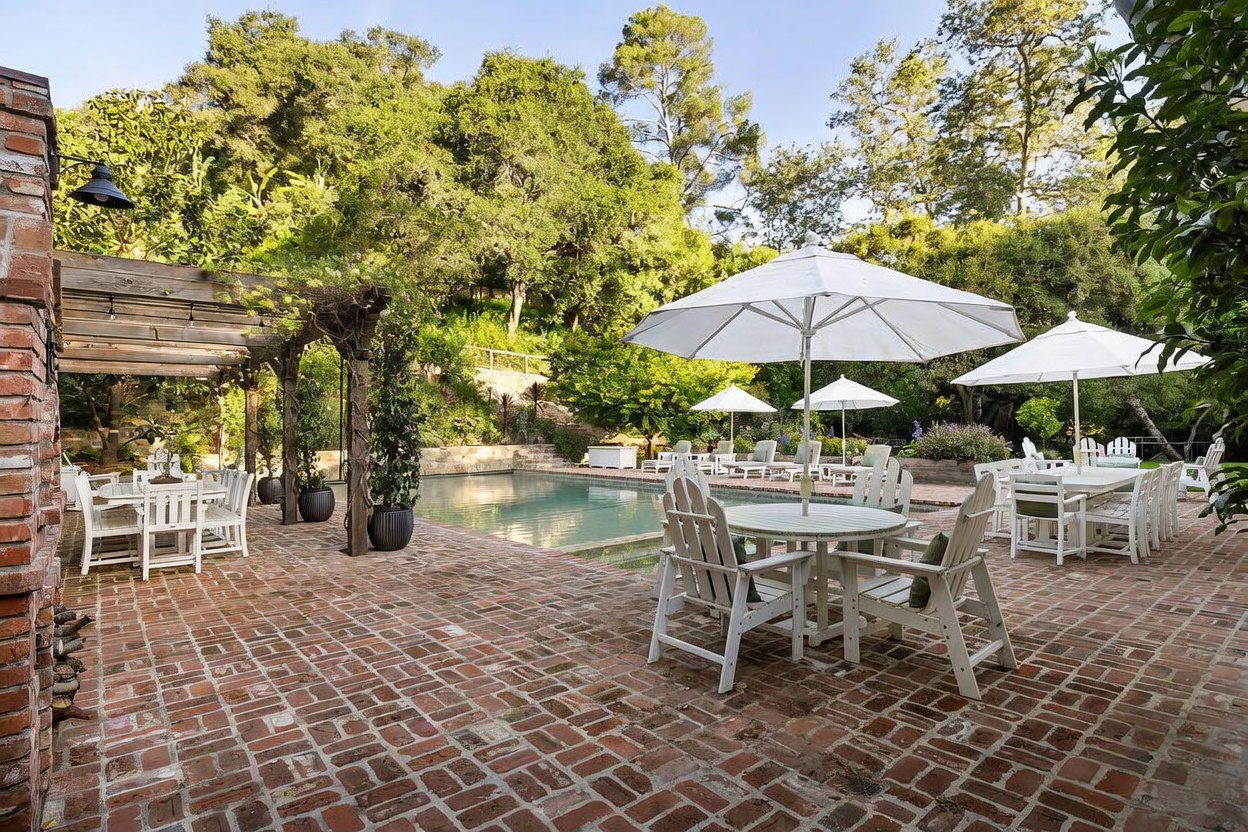 This expansive outdoor living area features a classic rectangular swimming pool surrounded by a spacious brick-paved patio. The space is furnished with multiple white dining sets under umbrellas and a pergola, creating an inviting environment for al fresco entertaining. Lush green trees and hillside landscaping provide a private, serene backdrop to this well-appointed backyard retreat.