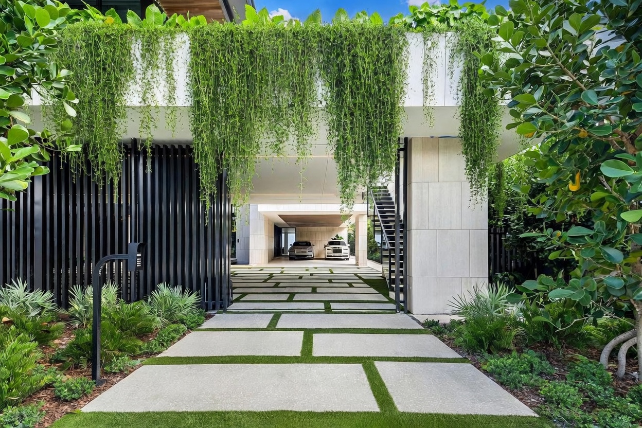 This modern entryway features a striking driveway paved with large concrete slabs separated by strips of lush green grass, leading toward a covered parking area. The structure is defined by clean, minimalist lines, vertical black slats, and a dramatic overhang draped with cascading greenery. The perspective is centered, drawing the eye through the open-air garage toward the vehicles parked in the distance.