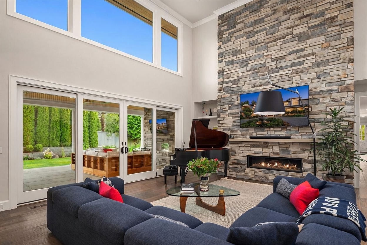 This spacious living room features a dramatic stacked-stone chimney breast that rises to the high ceiling, serving as a focal point above a linear fireplace. A grand piano sits near the center of the room, which is anchored by a large navy sectional sofa and a glass-topped coffee table. Tall glass sliding doors provide an expansive view of the outdoor patio and garden, filling the room with natural light.