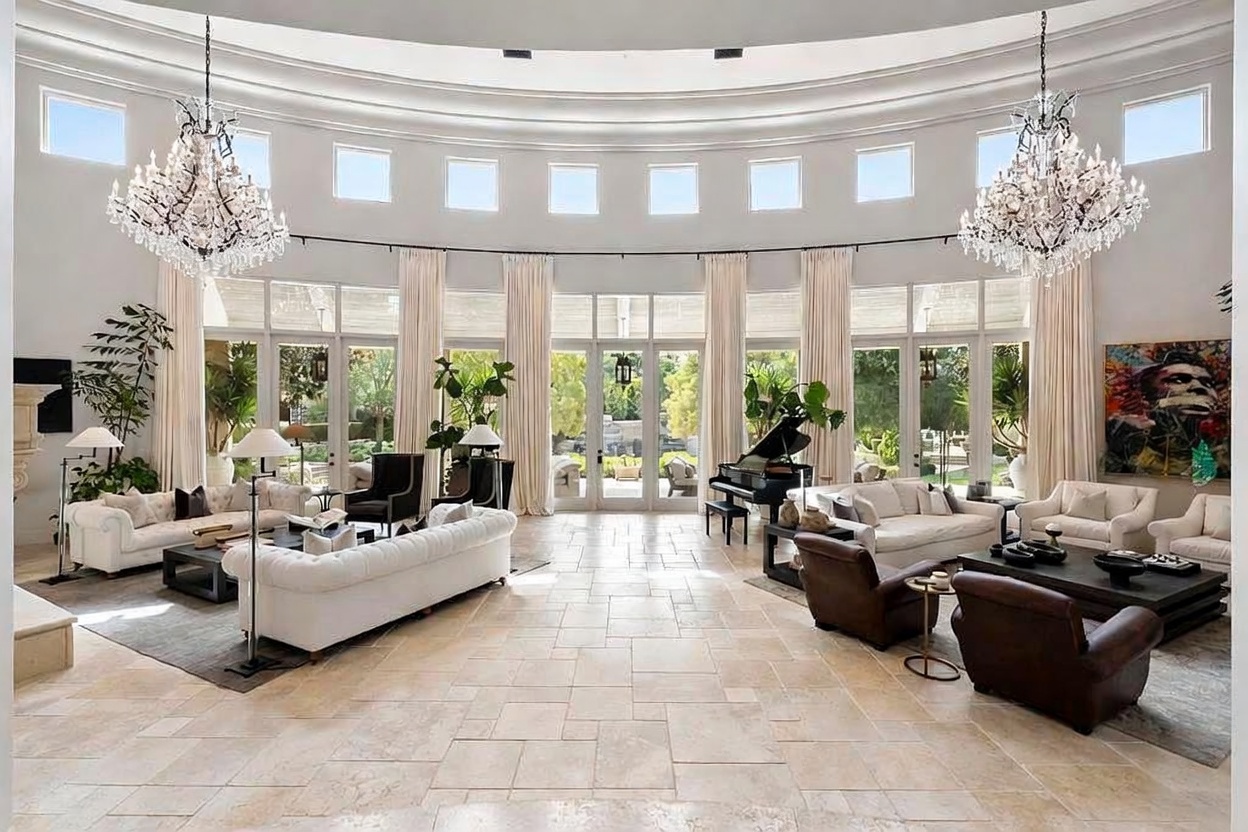 This is a grand living room featuring a curved wall of windows with elegant draperies, flooding the space with natural light. Two large crystal chandeliers hang from the high ceiling, adding a touch of luxury. The room is furnished with multiple white sofas, brown leather chairs, and a grand piano, creating a sophisticated and inviting atmosphere.