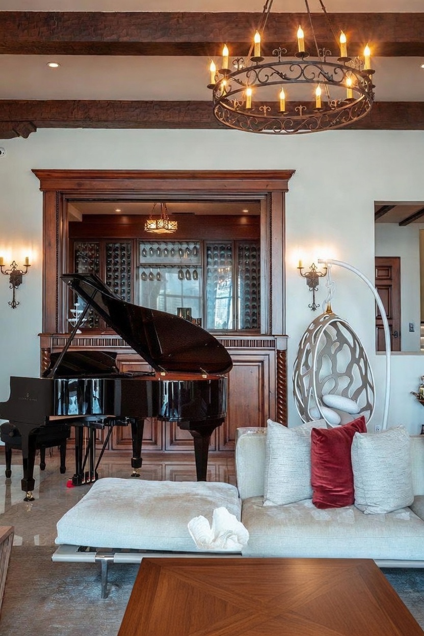 This sophisticated living space features a grand piano positioned in front of a custom wood-paneled wine display with glass doors. A modern, sculptural hanging chair sits beside a plush, light-toned sectional sofa, while a large, ornate iron chandelier hangs from a beamed ceiling. The room is finished with polished stone flooring and a coffee table adorned with a decorative floral arrangement, creating an atmosphere of refined luxury.