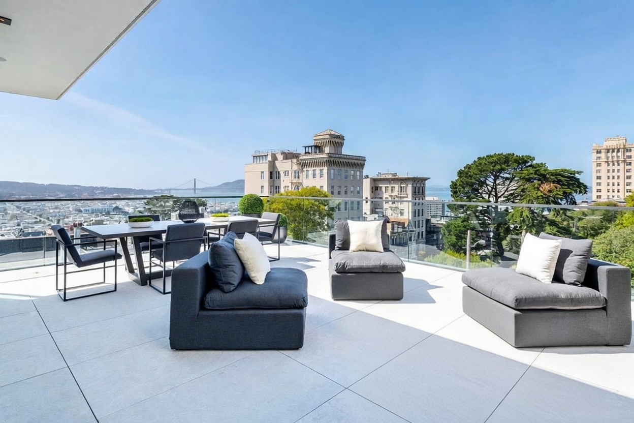 This expansive, modern terrace offers a sophisticated outdoor living space with panoramic city and bay views, including the Golden Gate Bridge. The area is furnished with a contemporary dining set and plush, dark-toned lounge seating, all arranged on light-colored stone pavers. A sleek glass railing provides an unobstructed perspective, creating a seamless transition between the elevated deck and the urban landscape beyond.