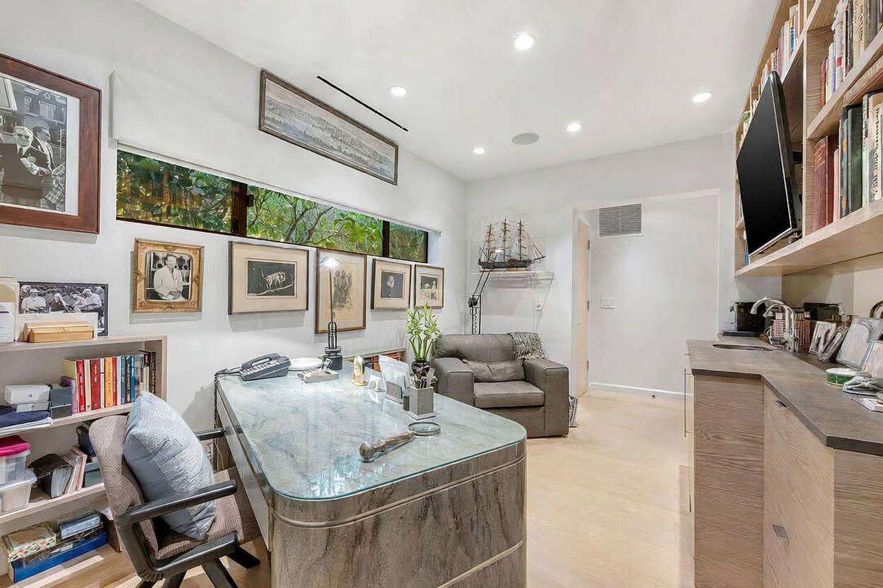 This well-appointed home office features a sleek, glass-topped desk with a textured base, positioned centrally in the room. The space includes built-in shelving filled with books, a comfortable armchair in the corner, and a wet bar area with cabinetry. A long, horizontal window provides natural light and a view of the outdoors, while framed artwork and a model ship add a personalized, sophisticated touch.