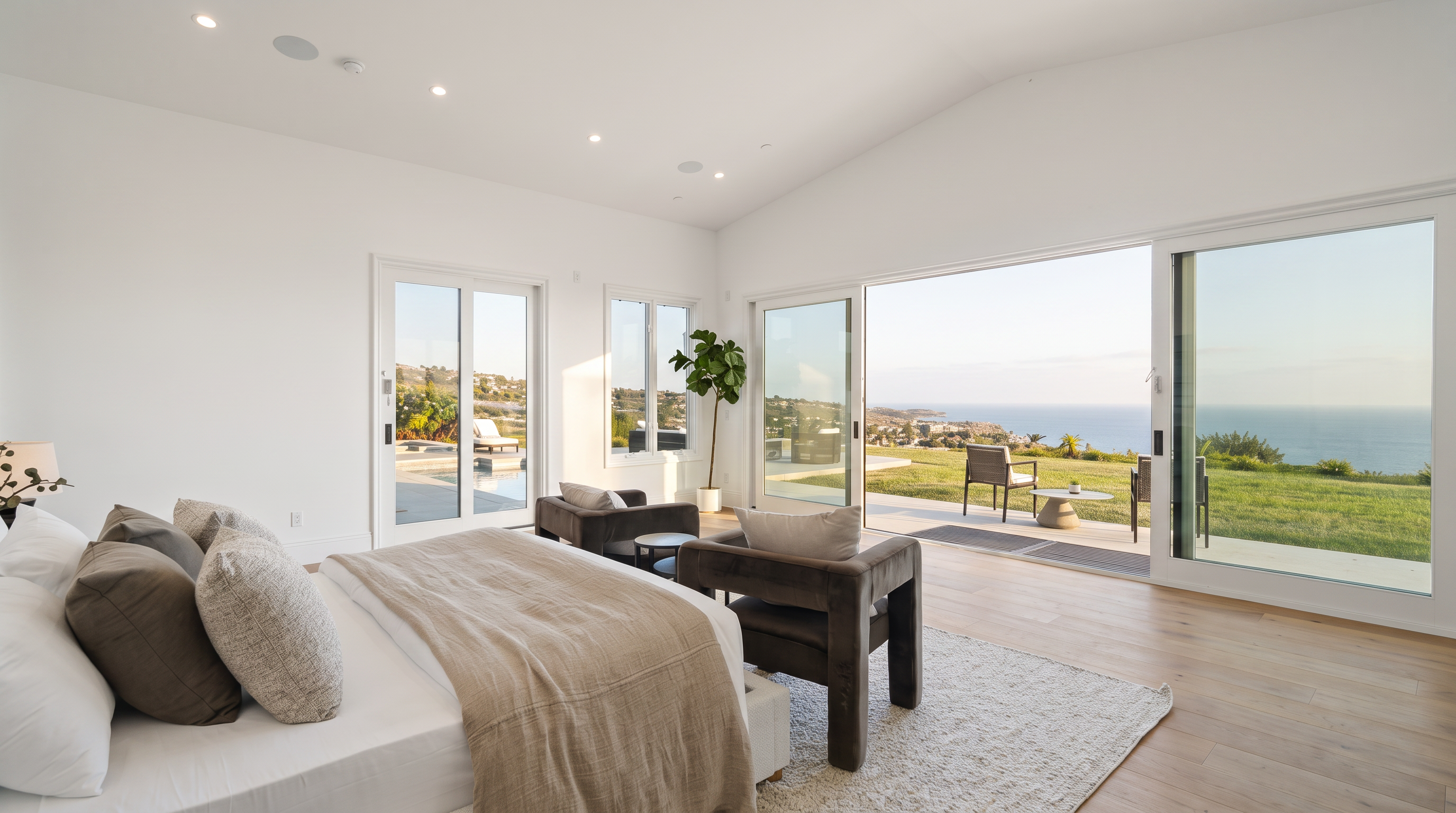 This spacious primary bedroom features a minimalist design with light wood flooring and a neutral color palette. Large sliding glass doors provide an expansive, unobstructed view of the ocean and coastline, seamlessly blending the indoor space with the outdoor patio. The room is furnished with a plush bed and two modern armchairs, creating a serene and luxurious atmosphere.