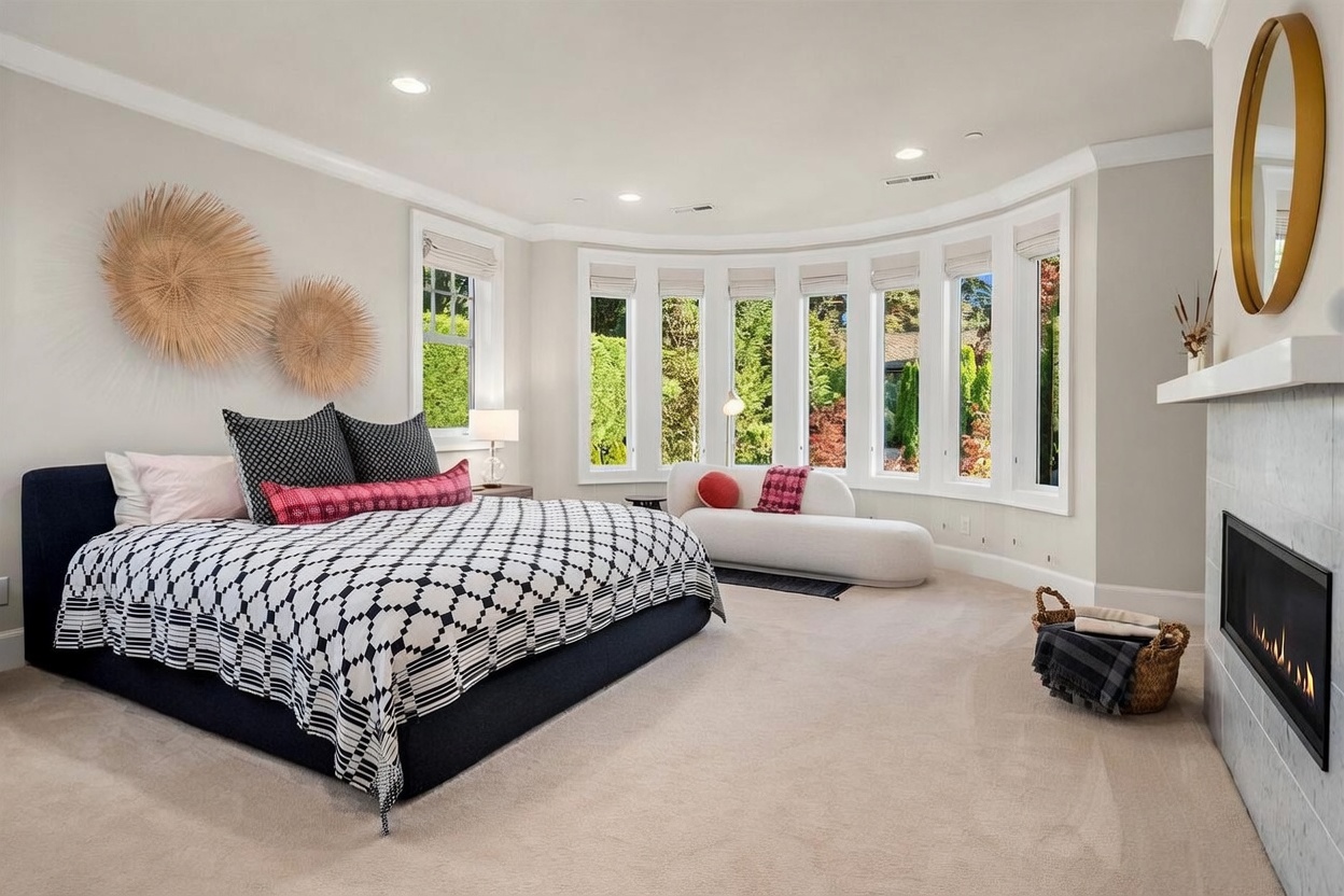 This spacious primary bedroom features a large bed with a dark upholstered frame and a bold black-and-white patterned quilt. A curved bay window area provides ample natural light and houses a modern, curved chaise lounge, while a fireplace with a light-colored surround adds a cozy focal point to the room. The space is decorated with textured wall art and a minimalist mirror, creating a sophisticated and serene atmosphere.