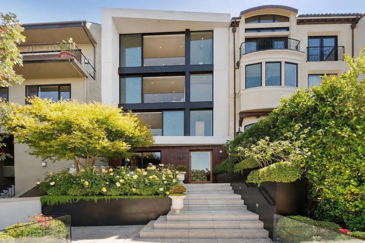 This modern, multi-story residence features a striking facade with large glass panels and a clean, minimalist aesthetic. A wide stone staircase leads up to the main entrance, flanked by lush landscaping, vibrant yellow roses, and a mature tree that adds a touch of natural warmth to the structure. The composition is captured from a low-angle, eye-level perspective, emphasizing the home's impressive scale and contemporary architectural design.