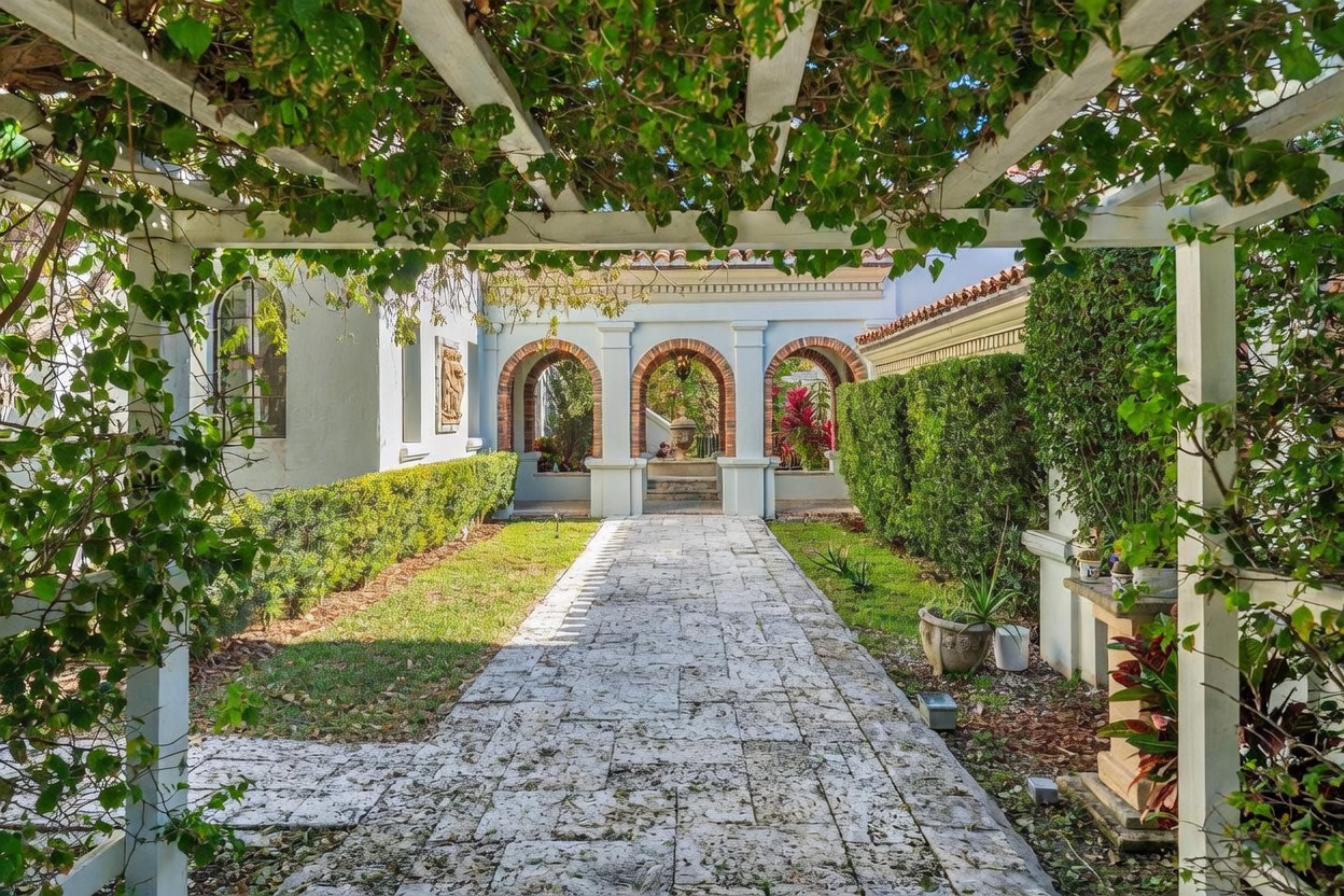 This serene garden walkway features a stone-paved path framed by a white wooden pergola draped in lush green vines. The path leads toward a series of elegant arched openings with brick detailing, which reveal a glimpse of a central fountain in the distance. Manicured hedges and vibrant landscaping create a private, tranquil atmosphere, evoking a Mediterranean-inspired aesthetic.
