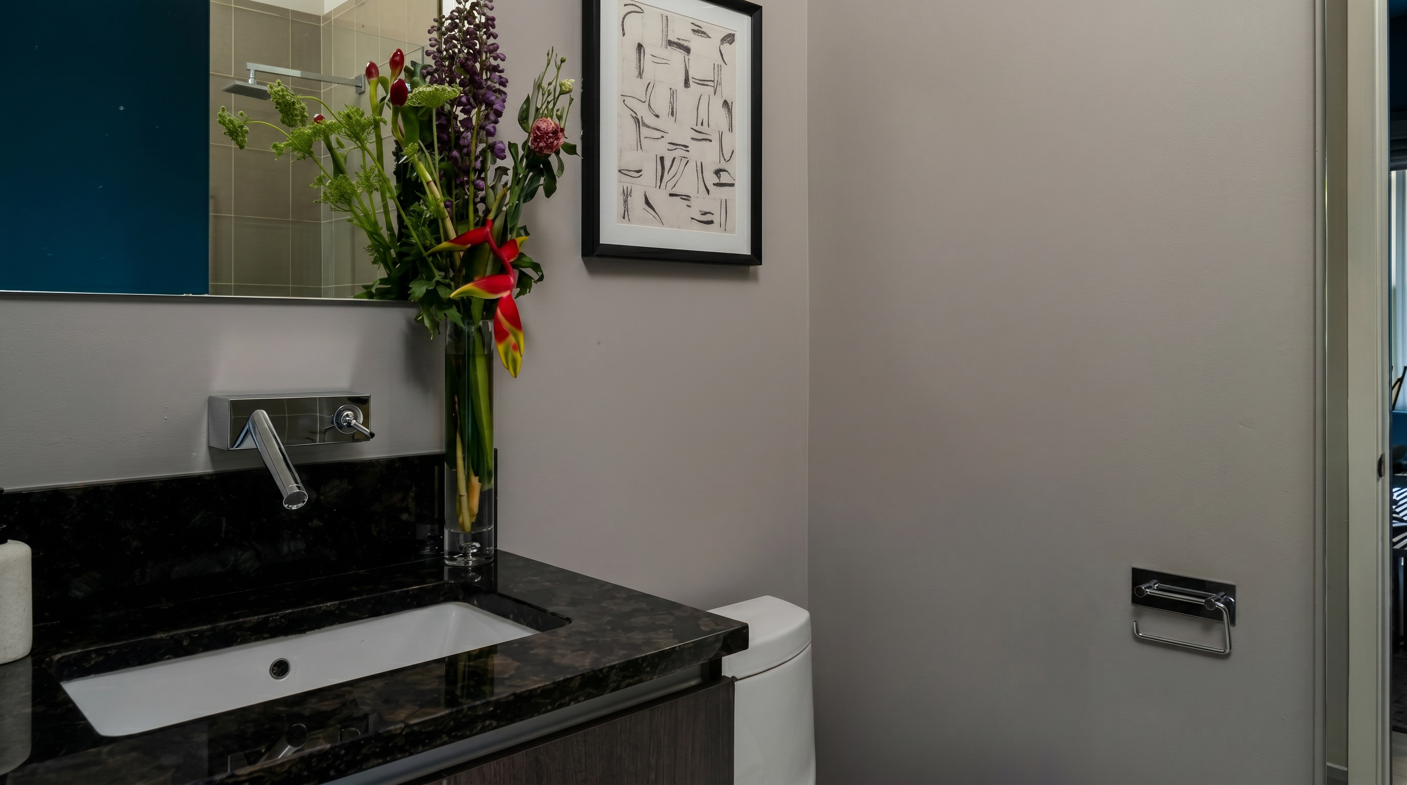 This compact powder room features a modern wall-mounted faucet over a sleek vanity with a dark marble countertop. The space is accented by a vibrant floral arrangement in a tall glass vase and a framed abstract art piece, creating a refined and polished aesthetic. A minimalist toilet and a chrome toilet paper holder complete the modern, functional design.