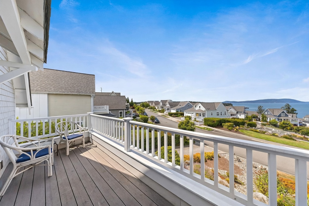 This elevated deck offers a serene outdoor living space with two white wicker chairs and a clear view of a quiet residential street and the distant coastline. The deck features composite decking and a white railing, providing a clean, coastal aesthetic. The perspective captures the expansive sky and the surrounding neighborhood, emphasizing the home's scenic location.
