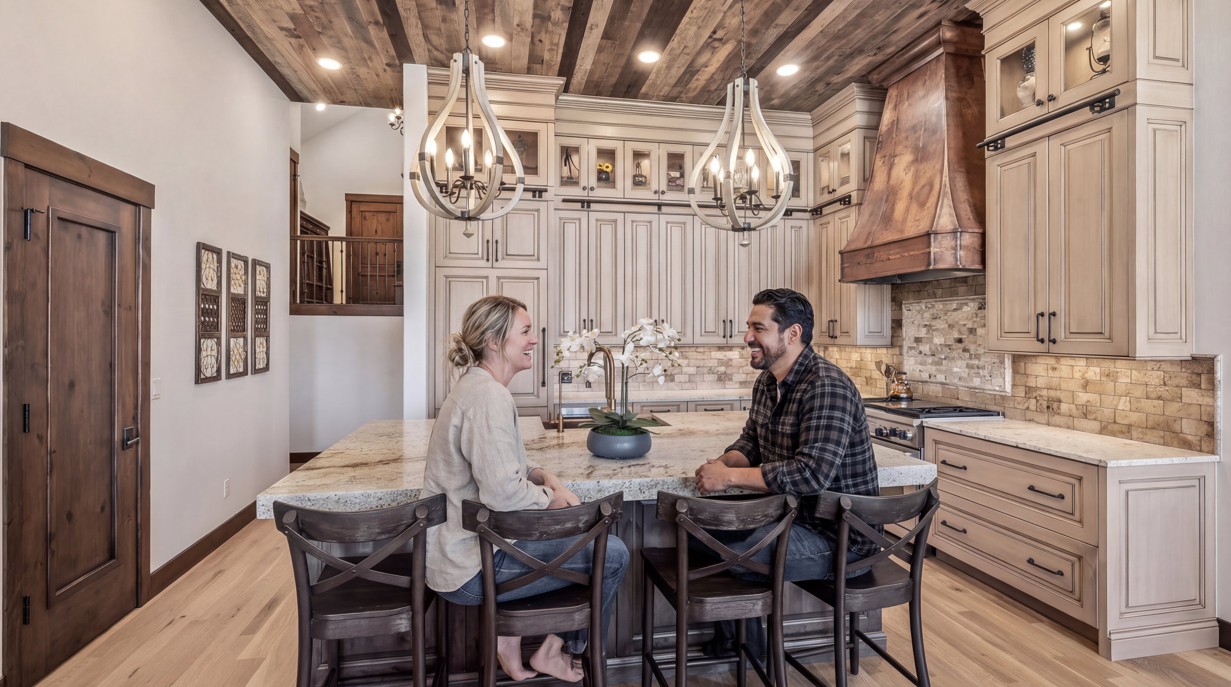 This stunning custom kitchen features a warm, rustic aesthetic with a unique wood-plank ceiling and a dramatic copper vent hood. The space is anchored by a spacious granite-topped center island, perfect for seating, and is complemented by light wood cabinetry with glass-front accents. A couple is shown gathered at the island, highlighting the home's inviting and open concept design for social living.