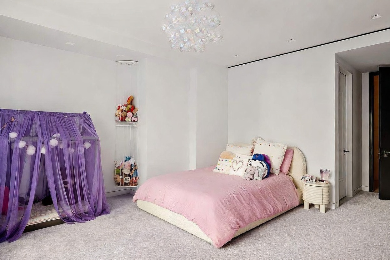 This bright and airy bedroom features a soft color palette with a plush pink bedspread and a whimsical purple canopy bed frame. A unique bubble-style chandelier hangs from the ceiling, while a vertical wall-mounted storage unit displays various stuffed animals. The room is finished with neutral carpeting and clean white walls, creating a playful and inviting atmosphere.