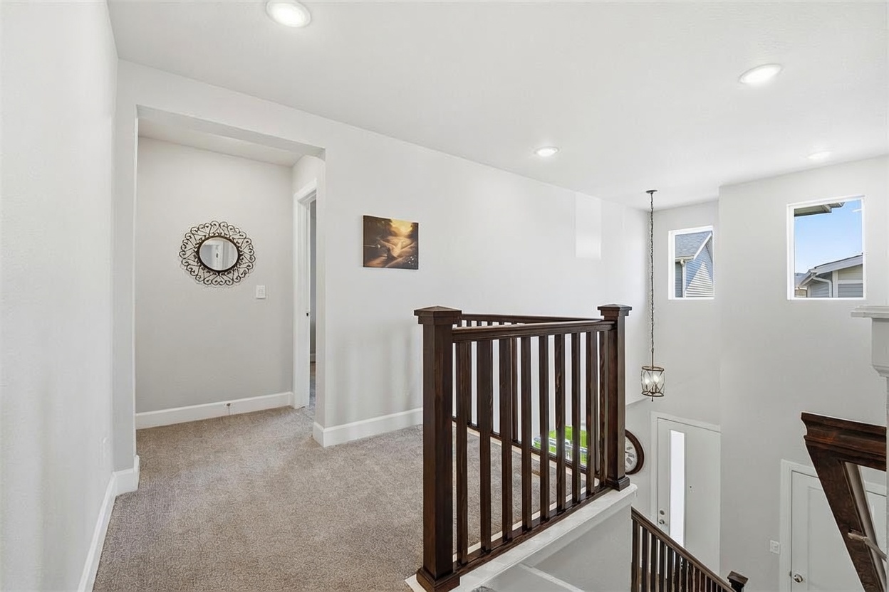 This image captures a bright, carpeted upper-level landing featuring a dark wood railing that overlooks the staircase below. The space is neutrally painted and includes a decorative circular wall mirror, a piece of wall art, and recessed lighting. The perspective provides a clear view of the transition between the hallway and the open stairwell, highlighting the home's airy and open floor plan.