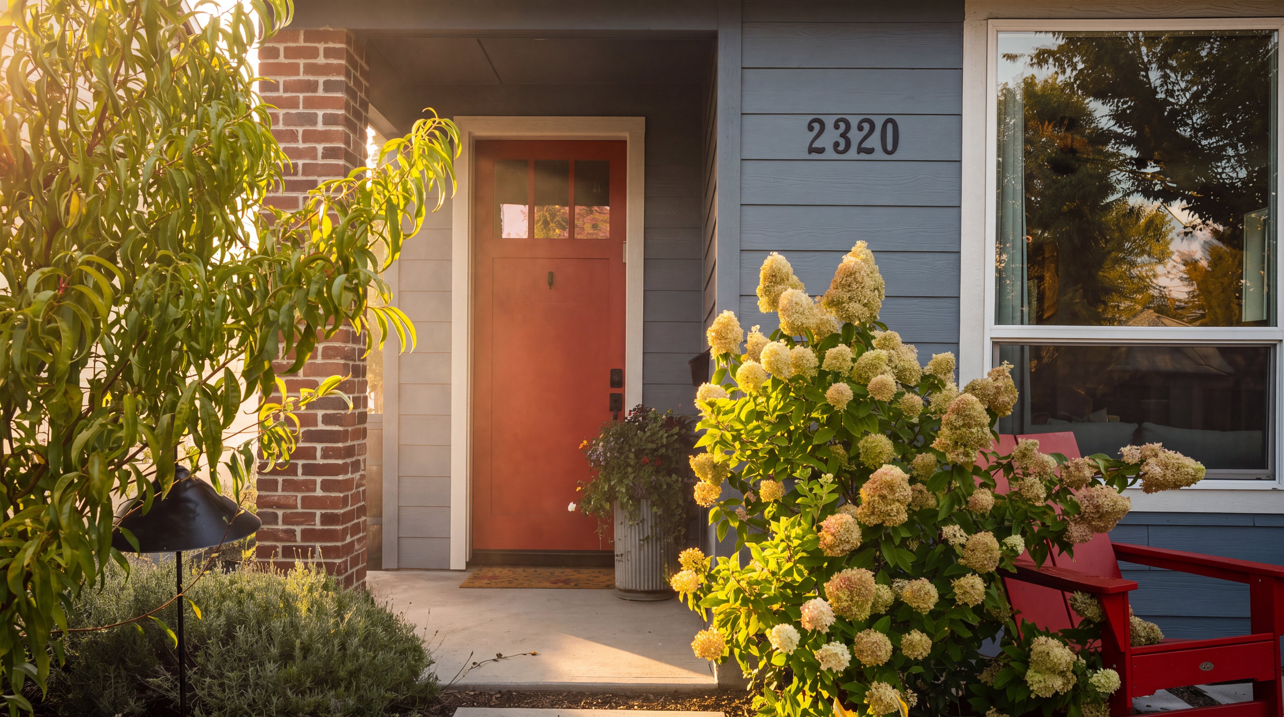 This inviting front entryway features a vibrant red door set against light blue horizontal siding and a brick-accented pillar. A lush, blooming hydrangea bush sits prominently in the foreground next to a red Adirondack chair, creating a welcoming and colorful curb appeal. The scene is captured from a ground-level perspective, highlighting the home's address number and the well-maintained landscaping.