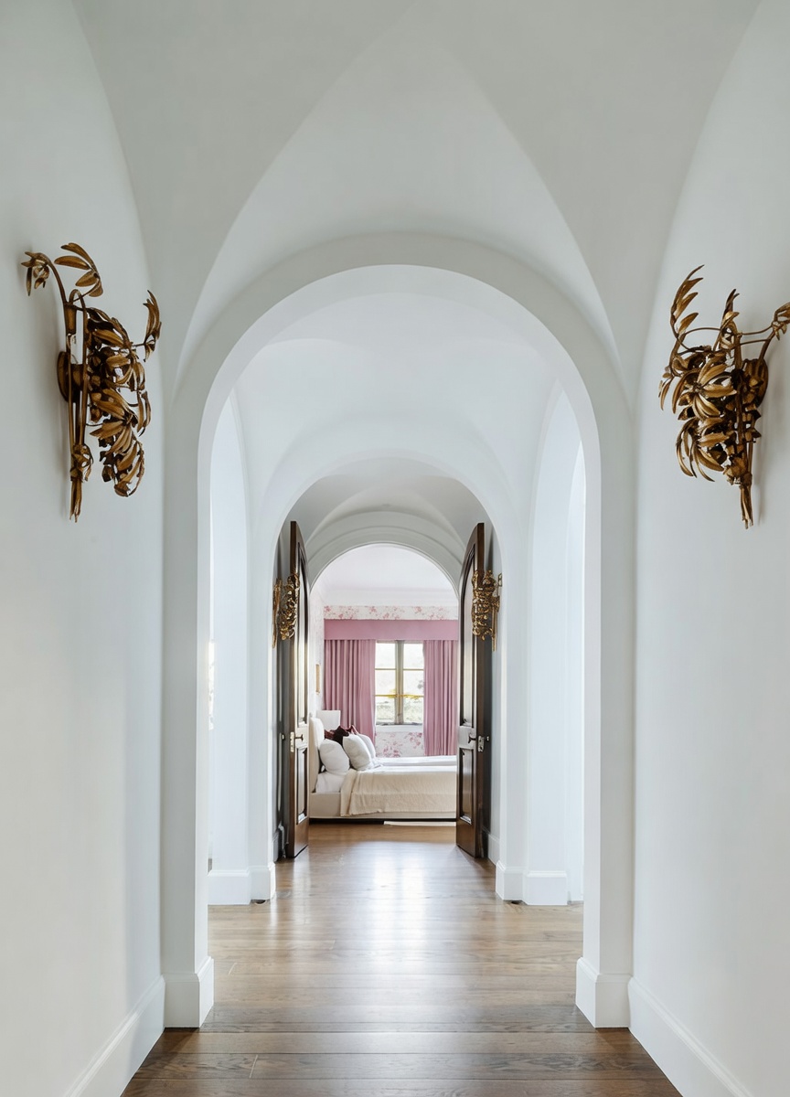 This elegant hallway features a series of dramatic, white-painted arched ceilings that create a sense of depth and architectural grandeur. The space is illuminated by ornate, gold-toned wall sconces with a leaf-like design, leading the eye toward a bedroom visible through an open doorway at the end of the hall. The flooring consists of warm-toned wood planks, providing a classic foundation for the bright, sophisticated interior.