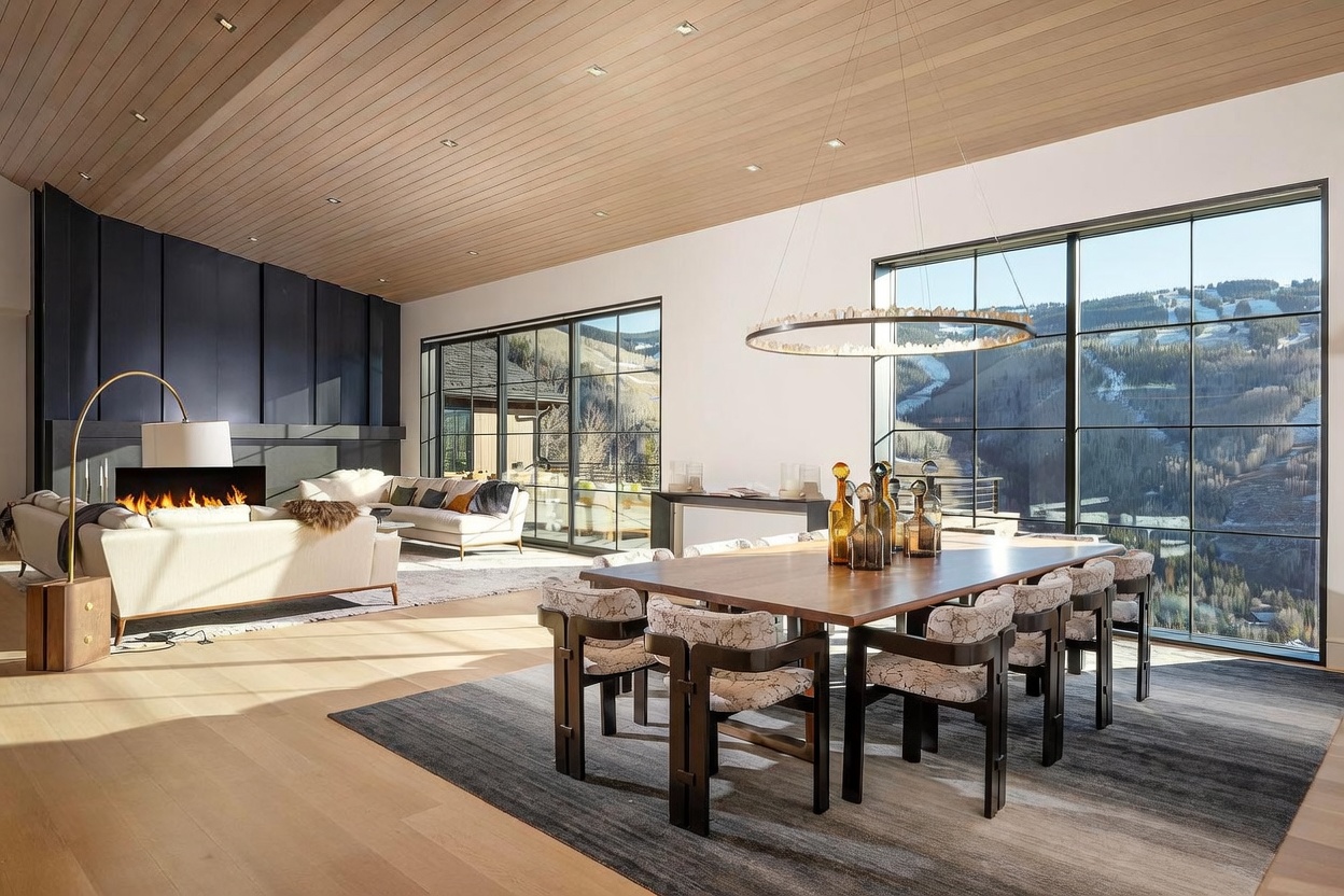 This expansive, open-concept living and dining area features a high, light-wood paneled ceiling and large windows that frame a scenic mountain view. The space is anchored by a contemporary fireplace with a dark, full-height surround and a comfortable seating arrangement, while a large dining table with patterned chairs sits under a circular chandelier. The room exudes a sophisticated, modern mountain aesthetic with a neutral color palette and clean architectural lines.