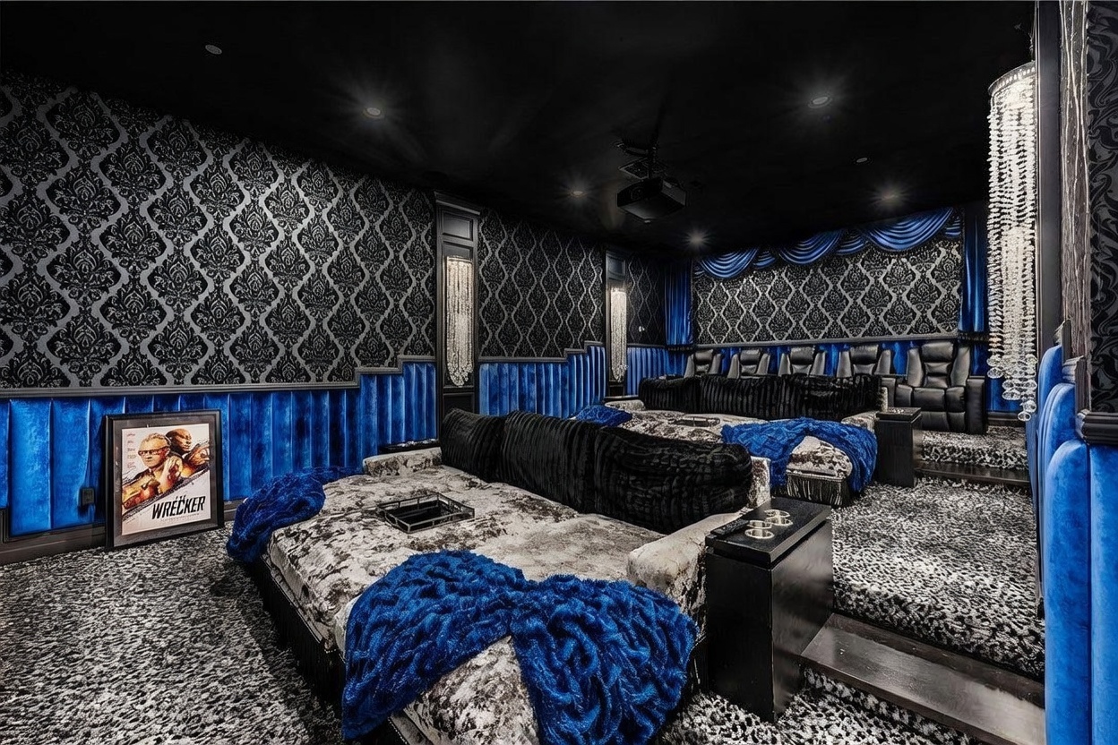 This opulent home theater features a dramatic, high-contrast design with deep blue velvet wall paneling and intricate black-and-white damask wallpaper. The tiered seating arrangement includes plush, velvet-upholstered loungers and a large, textured daybed, all accented by vibrant blue throws and crystal-adorned light fixtures. The room is designed for an immersive cinematic experience, complete with a projector mounted on the dark ceiling and a sophisticated, moody aesthetic.