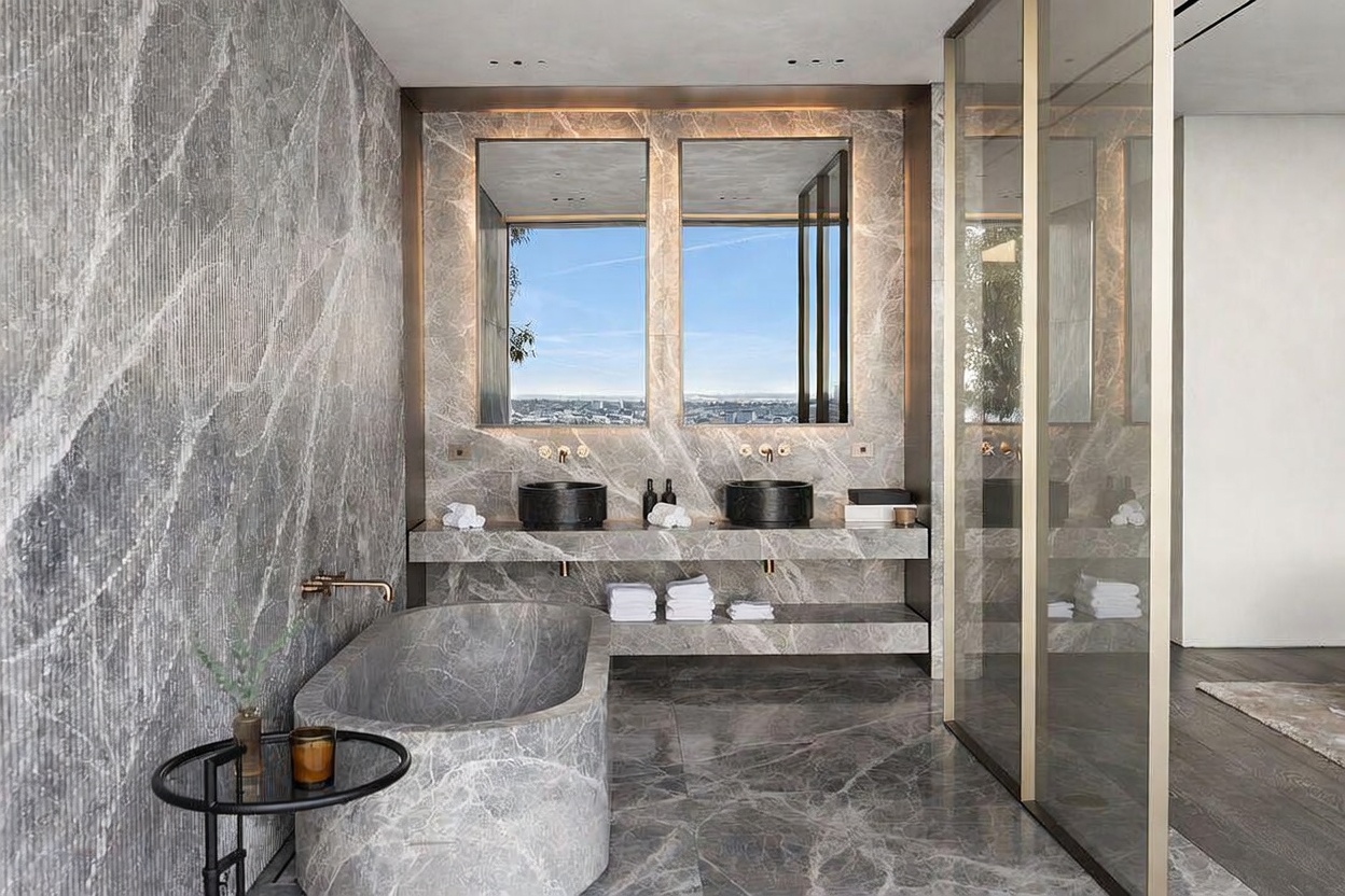 This luxurious primary bathroom features a sophisticated, monochromatic aesthetic dominated by high-end grey marble surfaces on the walls, vanity, and flooring. The space includes a deep, custom-carved stone soaking tub, a double-sink vanity with dark stone basins, and large windows that frame an expansive city view. The cinematic perspective highlights the seamless integration of natural stone textures and modern, minimalist design elements.