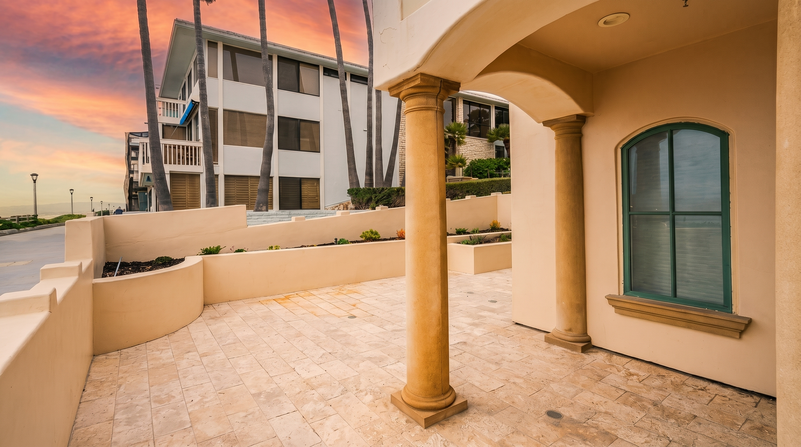 This image captures a covered outdoor patio area featuring light-toned stone tile flooring and two prominent columns supporting the structure above. The space opens up to a low-walled terrace with a small garden bed, offering a view of a multi-story building and a sunset sky in the background. The perspective is from under the covered porch, looking outward toward the exterior landscape.
