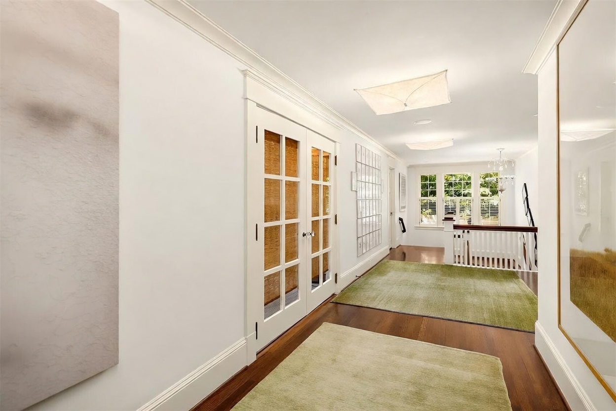 This bright and airy hallway features polished dark wood flooring accented by two light-colored area rugs. To the left, a set of white-framed French doors with textured glass panels leads into another room, while the right side opens to a staircase with a white railing and a view of a windowed landing. The space is illuminated by two square, flush-mount light fixtures on the ceiling, creating a clean and sophisticated transition area.