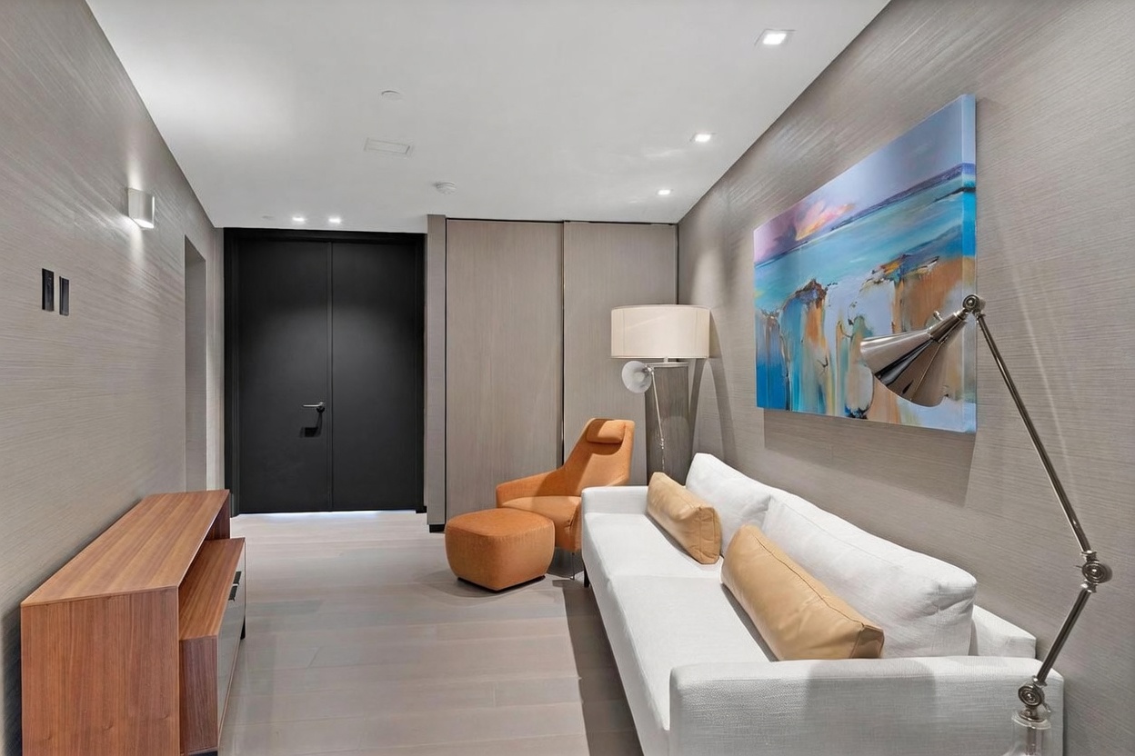 This contemporary living space features a white sofa accented with tan cushions, paired with a matching orange armchair and ottoman. The room is finished with textured wall coverings and light-toned wood flooring, while a large, vibrant abstract painting adds a pop of color to the wall. A sleek, modern floor lamp and a low-profile wooden media console complete the minimalist and sophisticated aesthetic.