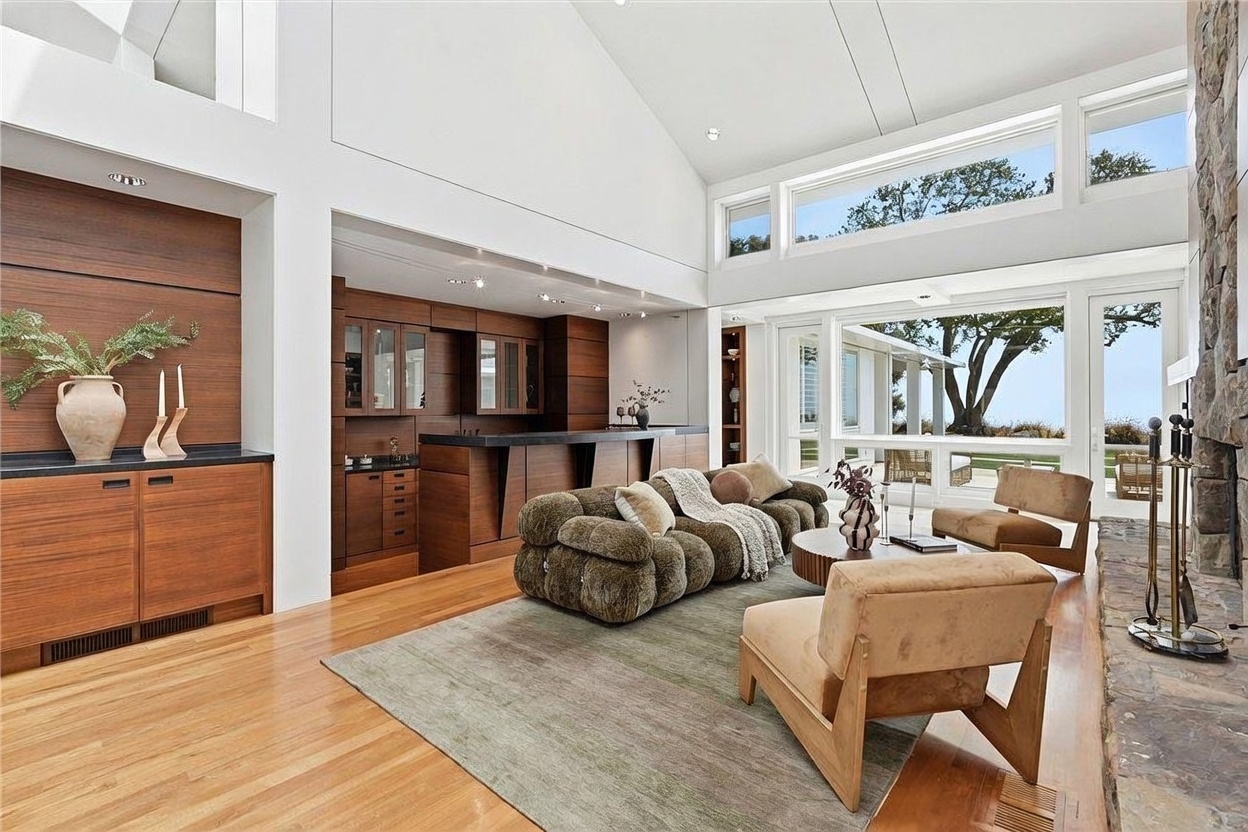 This spacious living room features a vaulted ceiling and a large, textured sofa centered on a neutral area rug. To the left, a built-in bar area with dark wood cabinetry provides a sophisticated focal point, while a stone-clad chimney breast anchors the right side of the room. Large windows and glass doors offer an expansive view of the outdoor landscape, filling the room with natural light.