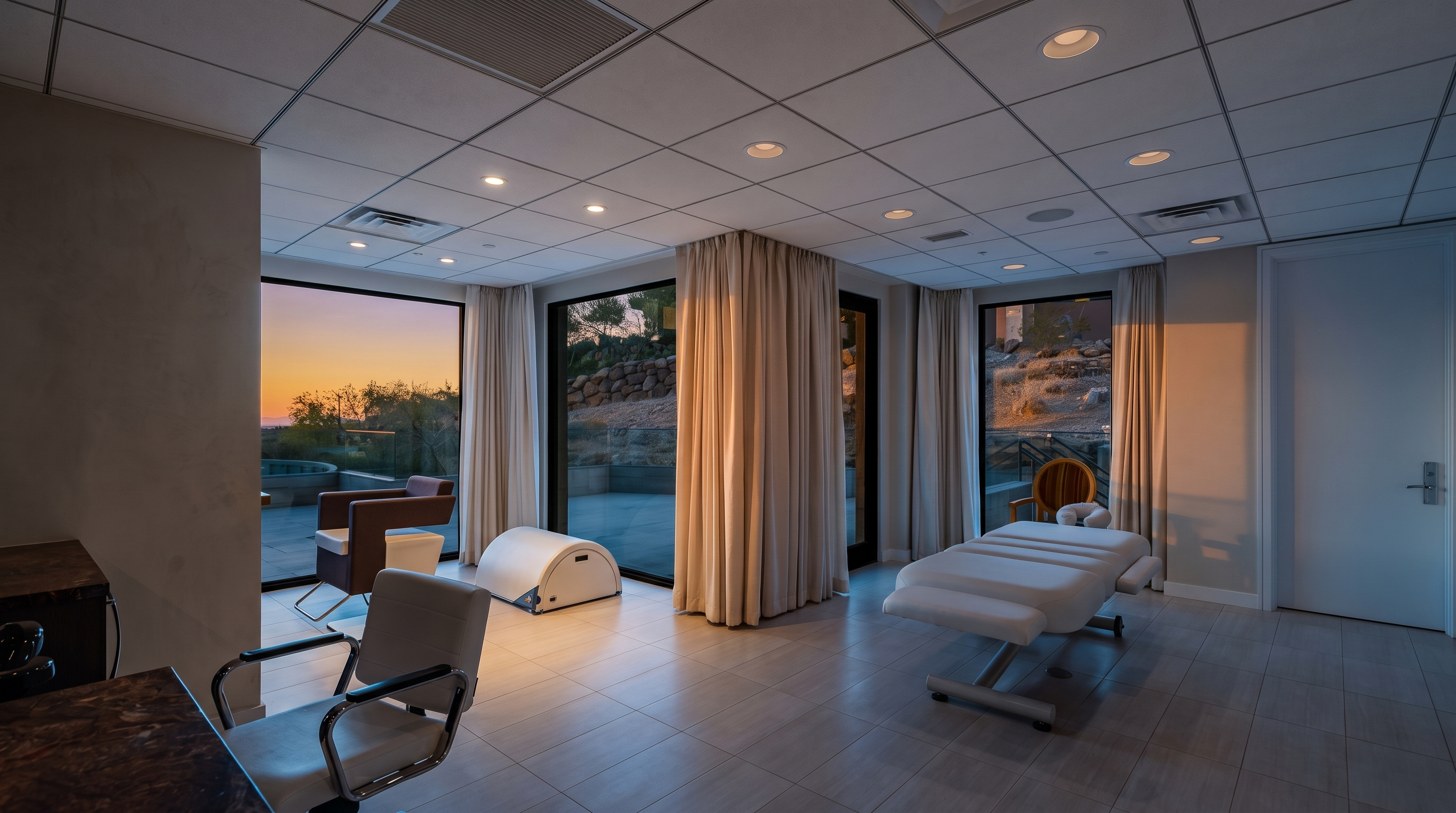 This bright, minimalist room appears to be a professional spa or massage treatment space, featuring a white massage table and a modern swivel chair. The room is characterized by large windows that offer a view of the outdoor landscape, complemented by neutral-toned curtains and light-colored tile flooring. The clean, clinical aesthetic is enhanced by recessed ceiling lighting and a neutral color palette, creating a serene and functional environment.