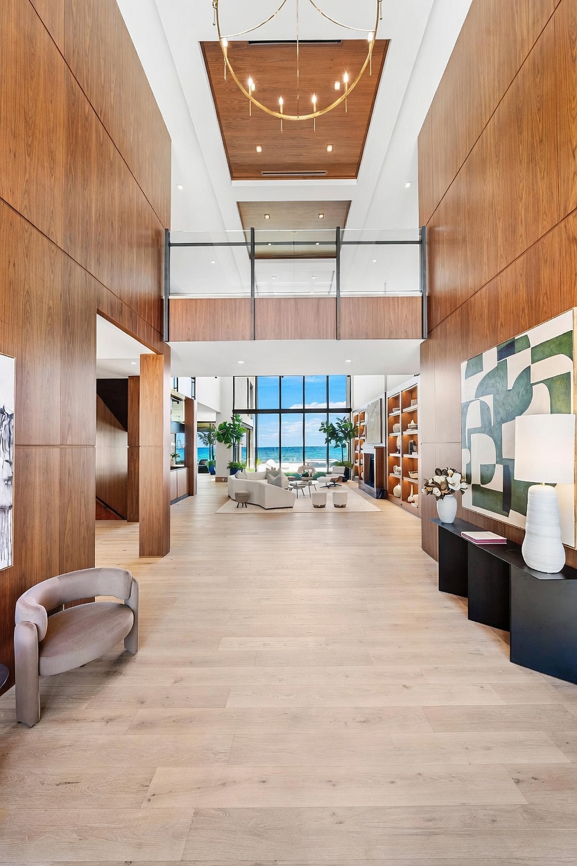 This grand entryway features soaring ceilings with wood-paneled accents and a large, circular chandelier. The space transitions into an open-concept living area with expansive glass walls that frame a clear view of the ocean. A sleek console table with a lamp and abstract art sits to the right, while a modern curved chair is positioned on the left, all set against light-toned wood flooring.