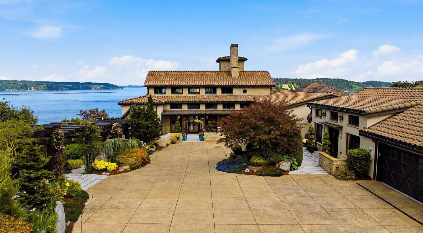 This expansive Mediterranean-style estate features a grand, paved motor court leading to a multi-level residence with a warm-toned tile roof and stone-clad exterior. The home is positioned overlooking a vast body of water, with lush landscaping, mature trees, and a pergola-covered walkway framing the entrance. The perspective is elevated, capturing the impressive scale of the property and its serene waterfront setting.