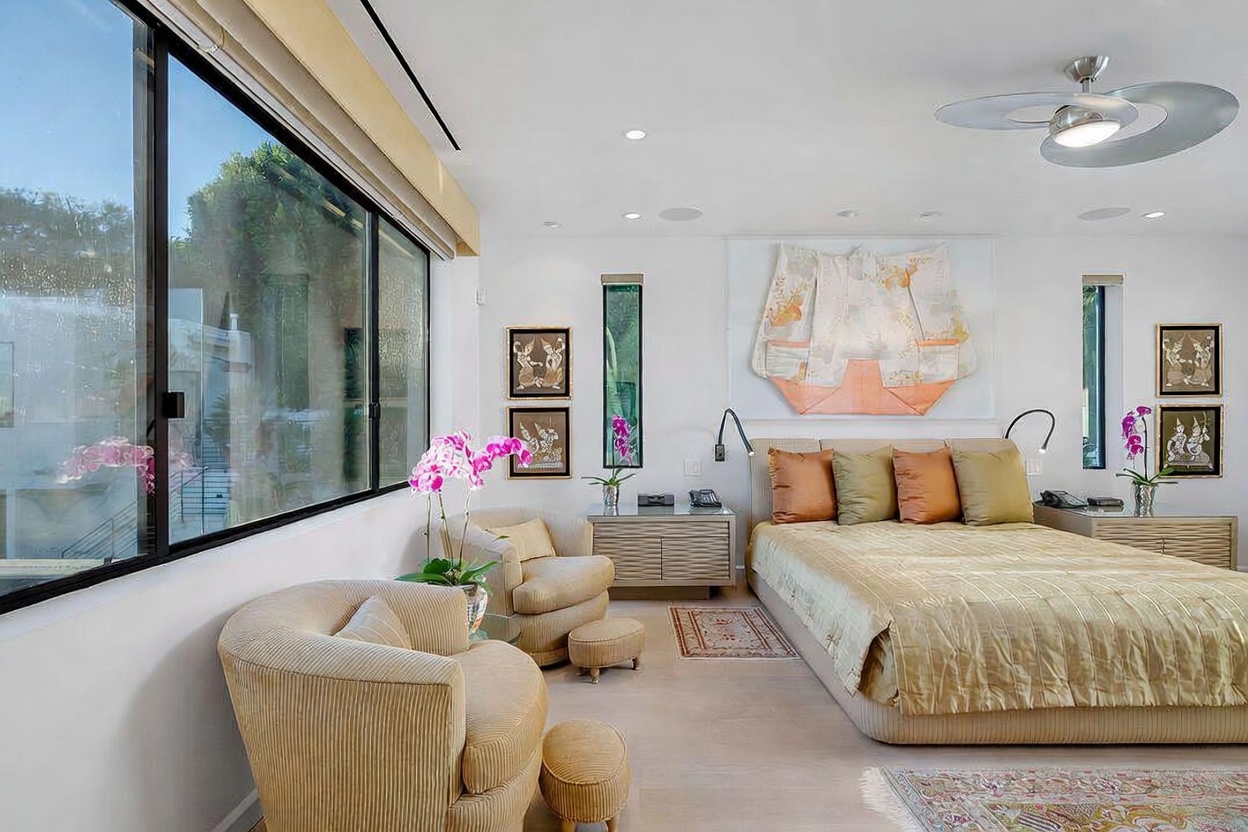 This spacious primary bedroom features a neutral color palette with soft textures and a large bed adorned with layered pillows. A wall of large windows allows for ample natural light, while decorative artwork and a unique wall-mounted textile piece add a sophisticated, curated aesthetic. The room is furnished with comfortable armchairs, matching nightstands, and a ceiling fan, creating a serene and inviting atmosphere.