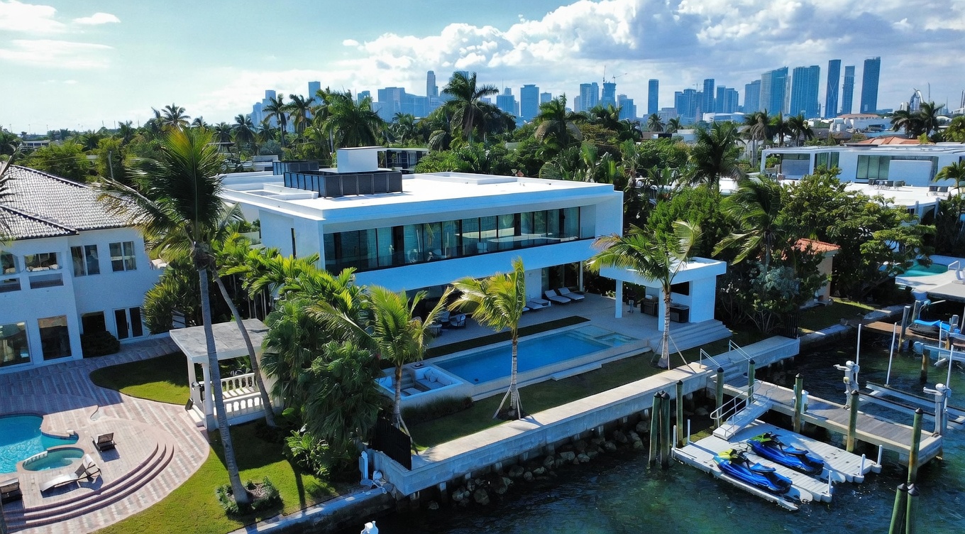 This aerial perspective captures a luxurious, modern waterfront estate featuring a sleek white architectural design with expansive glass walls. The property includes a private swimming pool, a covered outdoor lounge area, and a private dock equipped with jet skis on the canal. Lush tropical landscaping and palm trees surround the residence, with a panoramic view of the city skyline visible in the distance.