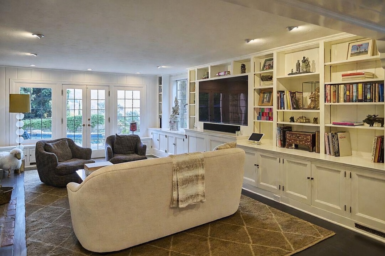 This bright and inviting living room features a large, cream-colored sofa facing a built-in wall unit that houses a television and decorative shelving. Two comfortable armchairs are positioned near French doors that offer a view of the outdoor pool area, creating a seamless indoor-outdoor flow. The space is anchored by a textured area rug and illuminated by recessed lighting, providing a warm and sophisticated atmosphere.
