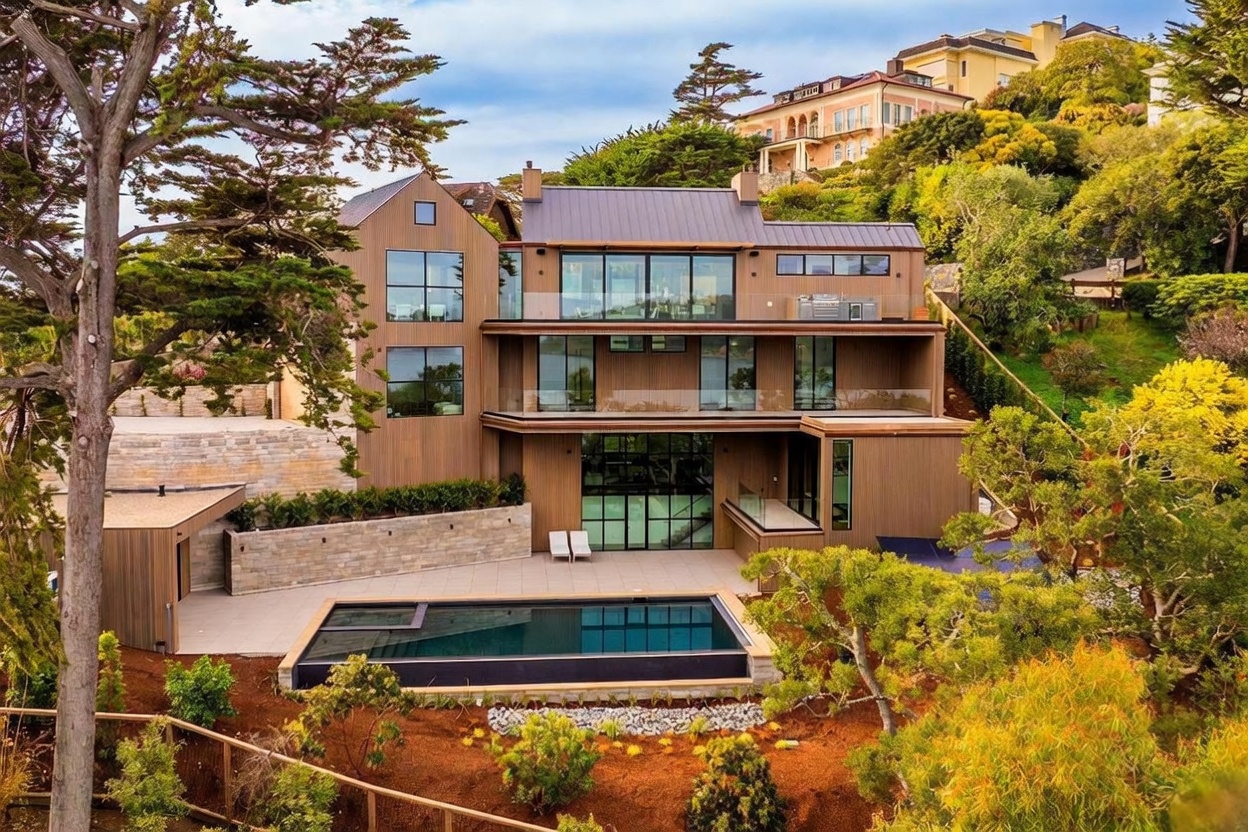 This striking multi-level modern residence is captured from an elevated rear perspective, showcasing its contemporary architectural design and integration with the hillside landscape. The home features extensive glass walls, multiple expansive balconies with glass railings, and a warm-toned wood-clad exterior. A sleek, rectangular infinity-edge pool sits on the lower terrace, complemented by stone retaining walls and lush, mature trees that frame the property.