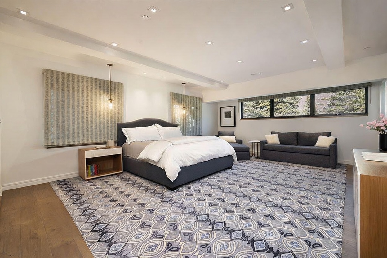 This spacious primary bedroom features a large bed with a dark upholstered headboard as the central focal point, flanked by light wood nightstands. The room is anchored by a bold, patterned area rug and includes a cozy seating area with a dark sofa and an armchair near the windows. The space is illuminated by recessed lighting and two pendant lights, creating a bright and inviting atmosphere.
