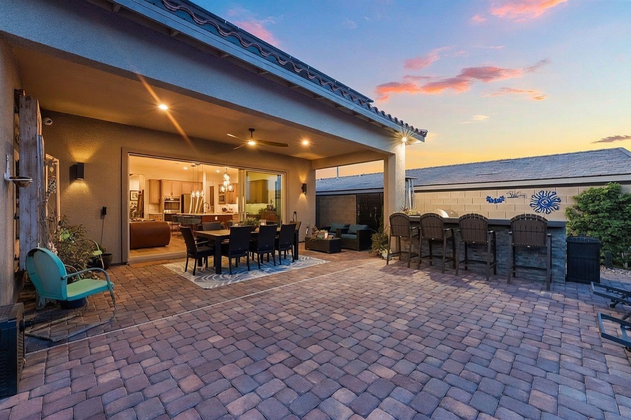 This spacious covered patio features a large dining area and a built-in outdoor bar, perfect for entertaining. The area is paved with stone pavers and overlooks a private backyard, with large sliding glass doors providing a seamless transition to the interior kitchen. The warm sunset lighting creates an inviting and relaxed atmosphere for outdoor living.