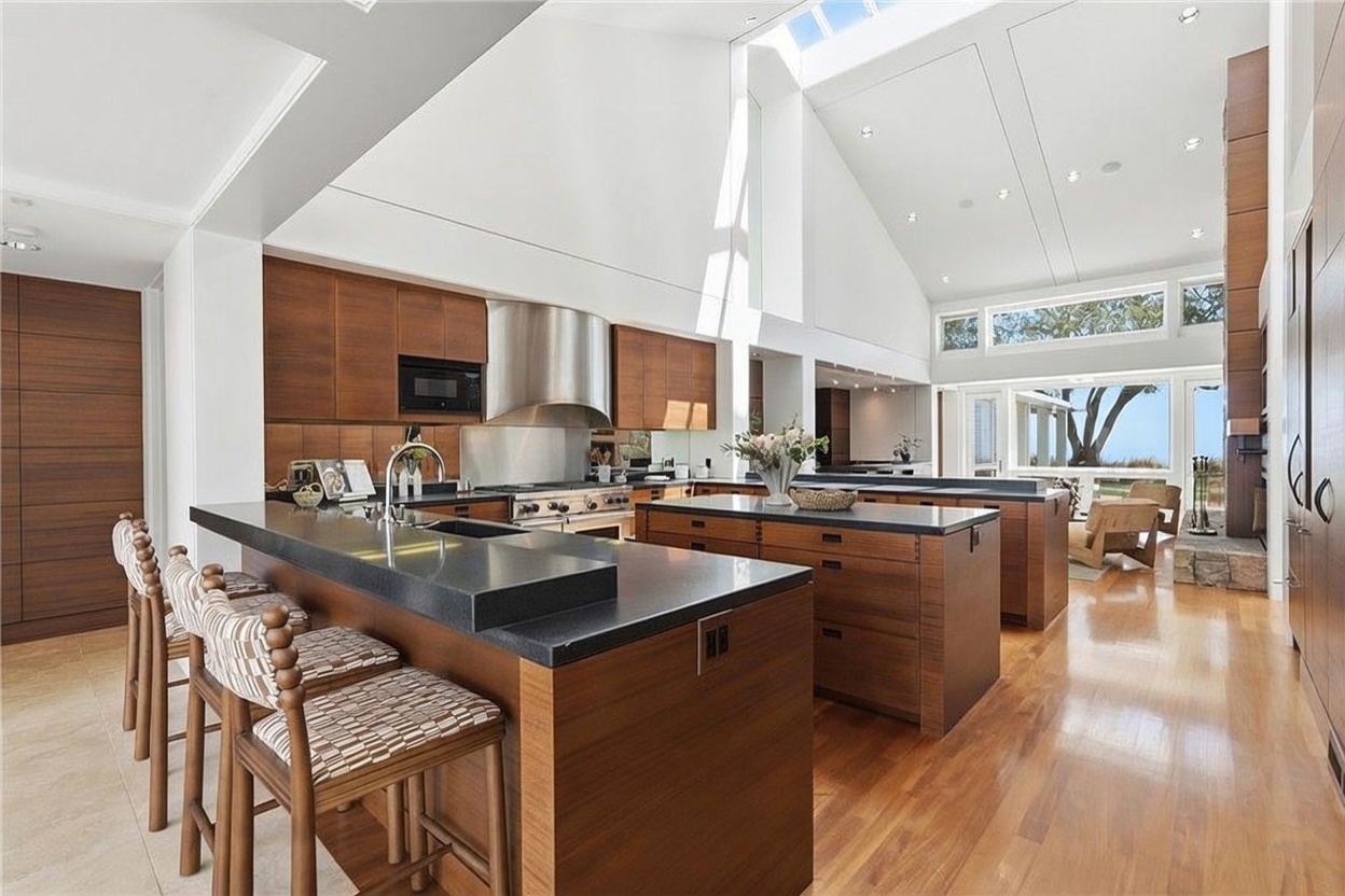 This expansive, high-ceilinged kitchen features warm wood cabinetry and a large central island with a dark stone countertop. The space is illuminated by natural light from large windows and a skylight, creating an airy and open atmosphere. A breakfast bar with patterned stools provides casual seating, while the clean lines and modern design offer a sophisticated, high-end aesthetic.