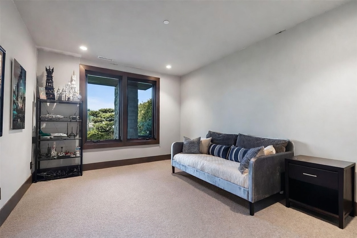 This versatile room features neutral walls, plush carpeting, and a comfortable sofa, making it suitable for a home office or a quiet sitting area. A tall shelving unit displays a collection of models, while large windows with dark wood trim provide a view of the outdoors. The space is well-lit by recessed lighting, creating a clean and inviting atmosphere.