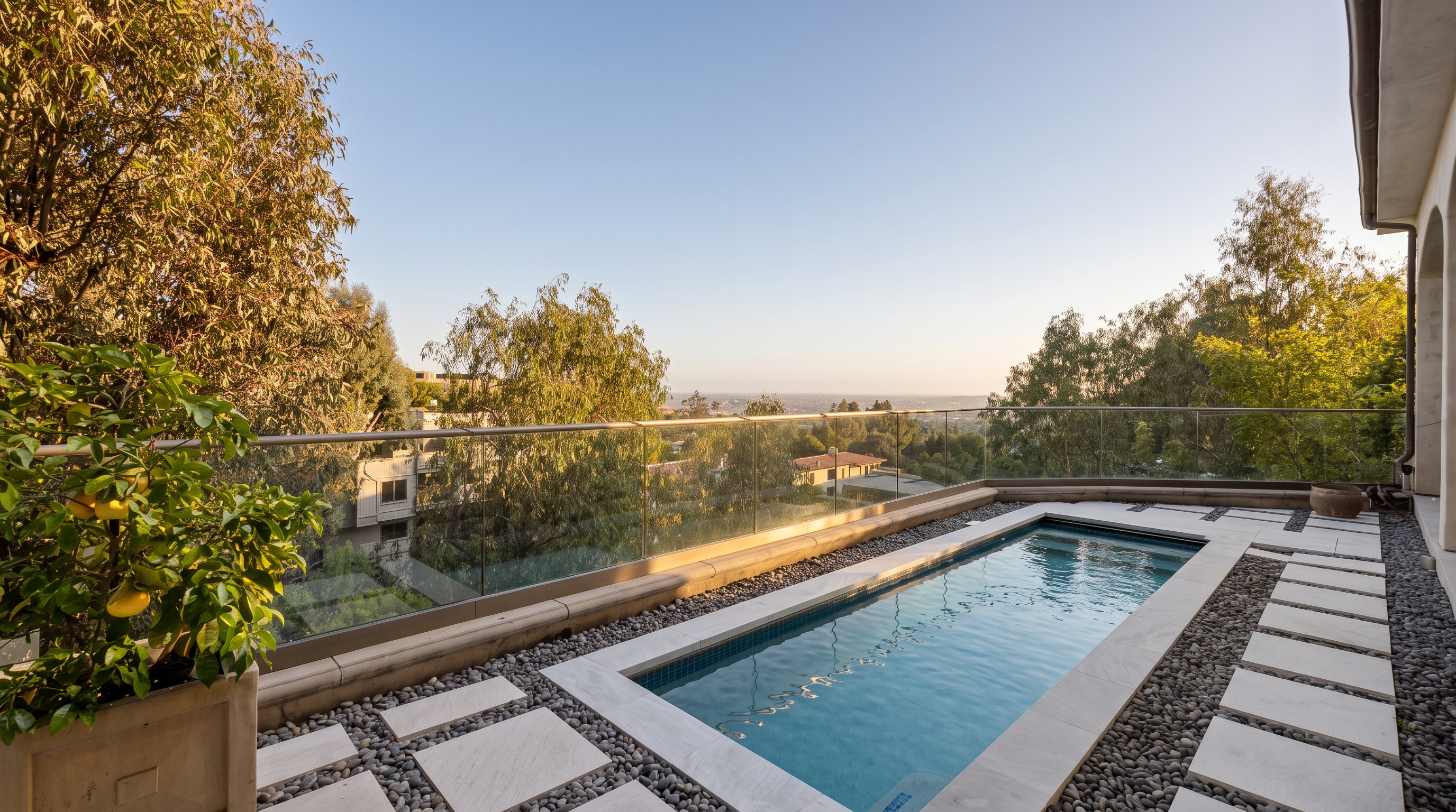 This elevated terrace features a sleek, rectangular plunge pool set into a stone deck surrounded by decorative river rocks. A glass railing provides an unobstructed, panoramic view of the surrounding landscape and distant horizon. The space is accented by a large potted lemon tree, creating a serene and luxurious outdoor retreat.