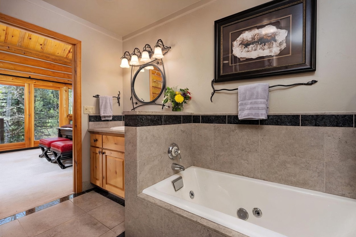 This primary bathroom features a jetted soaking tub with neutral stone tile surround and a separate vanity area with light wood cabinetry. A decorative wrought-iron light fixture hangs above a circular mirror, while a framed nature-themed artwork adds a rustic touch to the wall. The space opens into an adjacent room with large windows, creating a bright and airy transition between the bathroom and the living area.