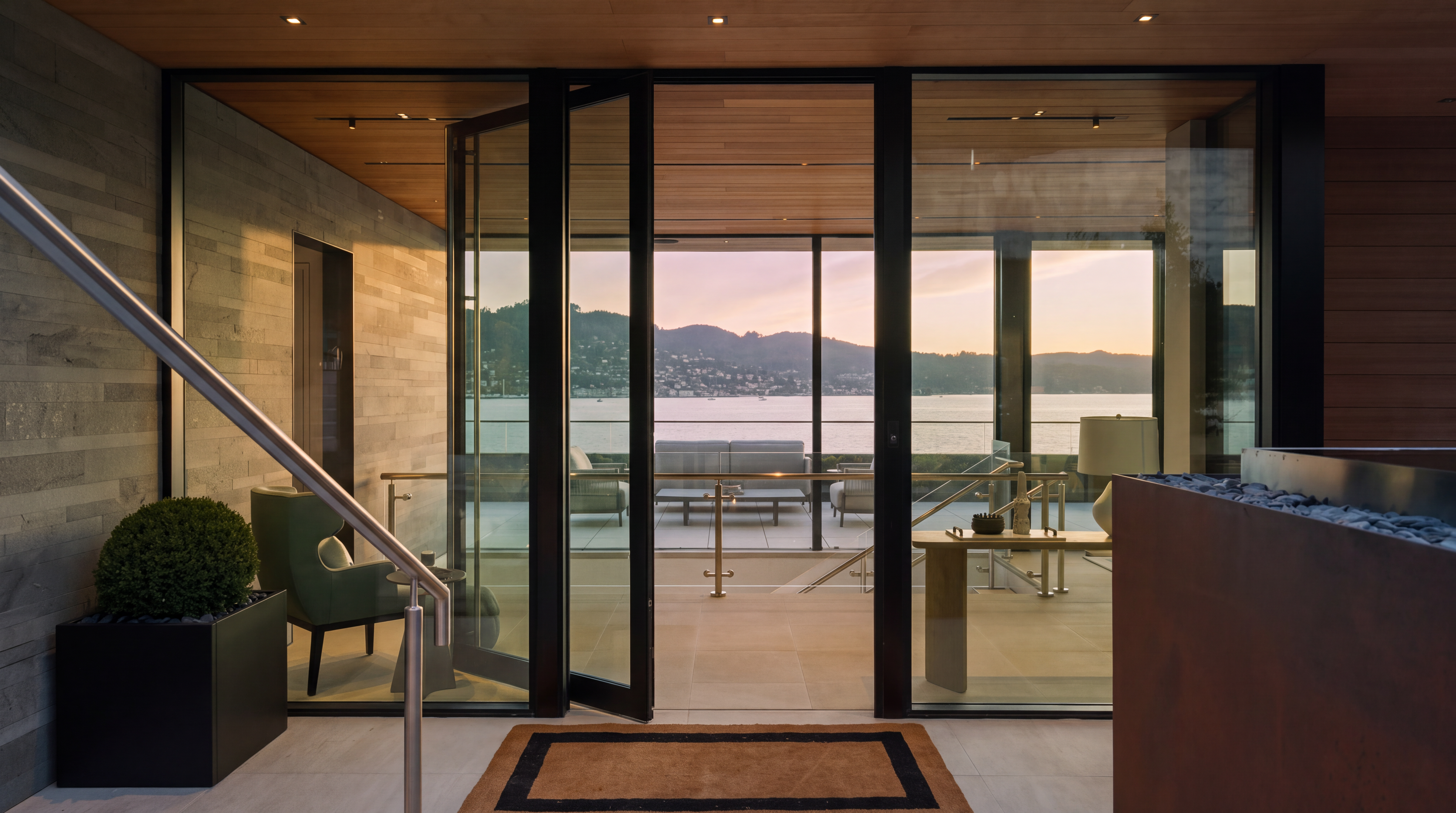 This sophisticated entryway features a modern aesthetic with a warm wood-paneled ceiling and a striking, illuminated handrail leading toward a glass-paneled door. Through the open doorway, a seamless transition reveals a contemporary living space that opens onto a deck overlooking a serene bay at sunset. The combination of natural stone wall textures, clean lines, and the dramatic golden-hour light creates an inviting and luxurious atmosphere.