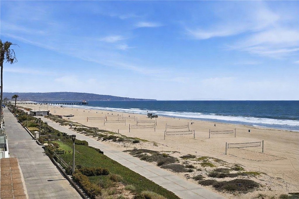 This elevated view from a balcony captures a stunning, expansive panorama of a sandy beach and the Pacific Ocean. The scene features a paved walkway running parallel to the shoreline, dotted with numerous volleyball nets set up on the sand, and a distant pier extending into the water. The bright, clear blue sky and the rhythmic waves create a serene and highly desirable coastal atmosphere.