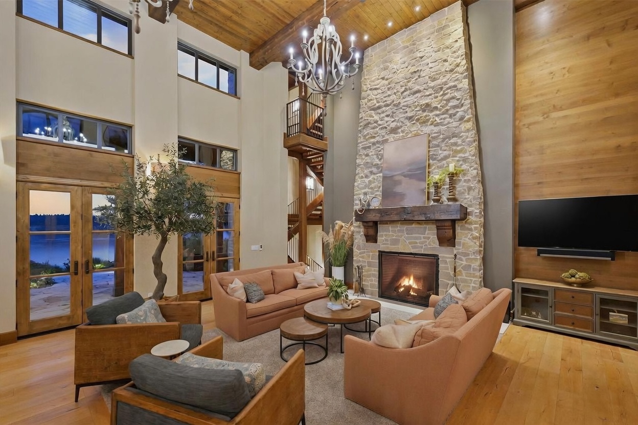 This expansive living room features a soaring vaulted ceiling with exposed wood beams and a prominent stone-clad chimney breast rising above the mantel. The space is furnished with two plush sofas and two armchairs arranged around a set of round coffee tables, all set upon a large area rug. Large windows and glass doors provide abundant natural light and views of the outdoors, while a grand chandelier serves as a focal point in the center of the room.