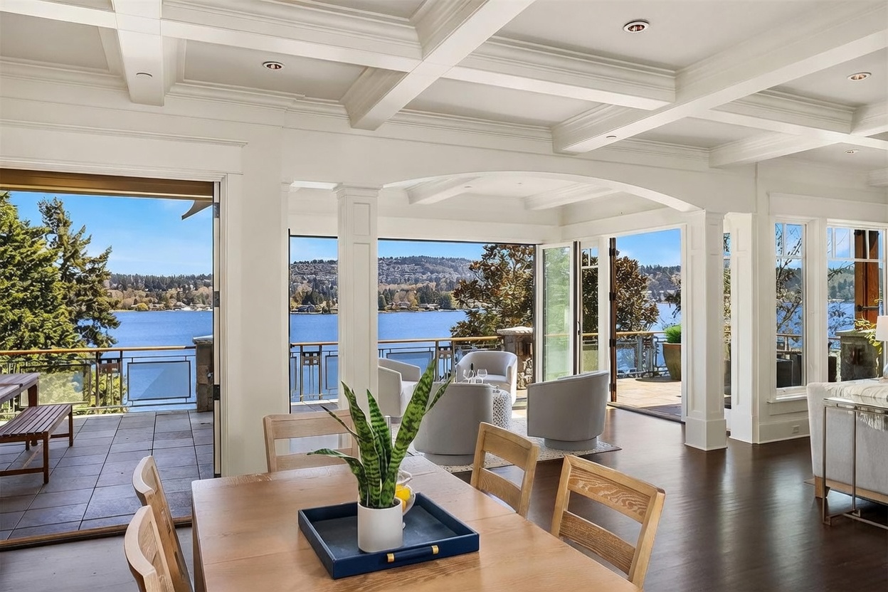 This elegant, open-concept living and dining area features a coffered ceiling and expansive floor-to-ceiling glass doors that seamlessly blend the indoor space with a scenic waterfront deck. The room is furnished with contemporary, neutral-toned seating and a wooden dining table, creating a sophisticated atmosphere that highlights the panoramic lake views. The perspective captures the transition from the refined interior to the serene outdoor environment, emphasizing the home's luxury and connection to nature.