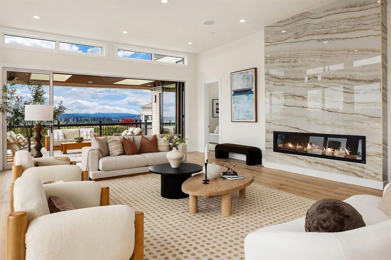 This bright and airy living room features a prominent stone-clad chimney breast with a wide, linear fireplace as its focal point. The space is furnished with plush, neutral-toned seating arranged on a patterned area rug, creating a comfortable and sophisticated atmosphere. Large sliding glass doors open to a covered outdoor patio, seamlessly blending the indoor living area with the scenic landscape beyond.