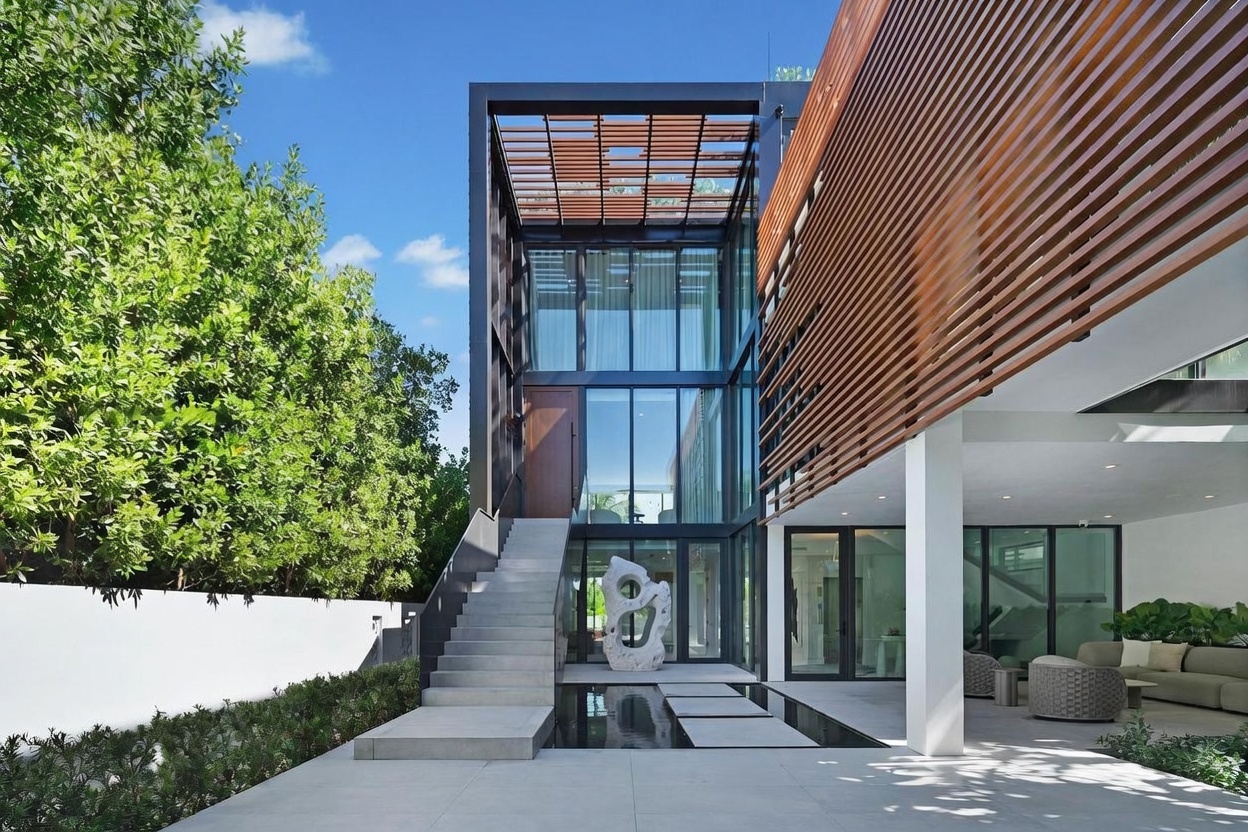 This striking modern entryway features a dramatic glass-enclosed staircase and a prominent wooden front door. A concrete staircase leads up to the entrance, positioned above a reflective water feature with stone stepping stones. The exterior combines clean white walls, dark metal framing, and warm wood-slat accents, creating a sophisticated and architectural aesthetic.