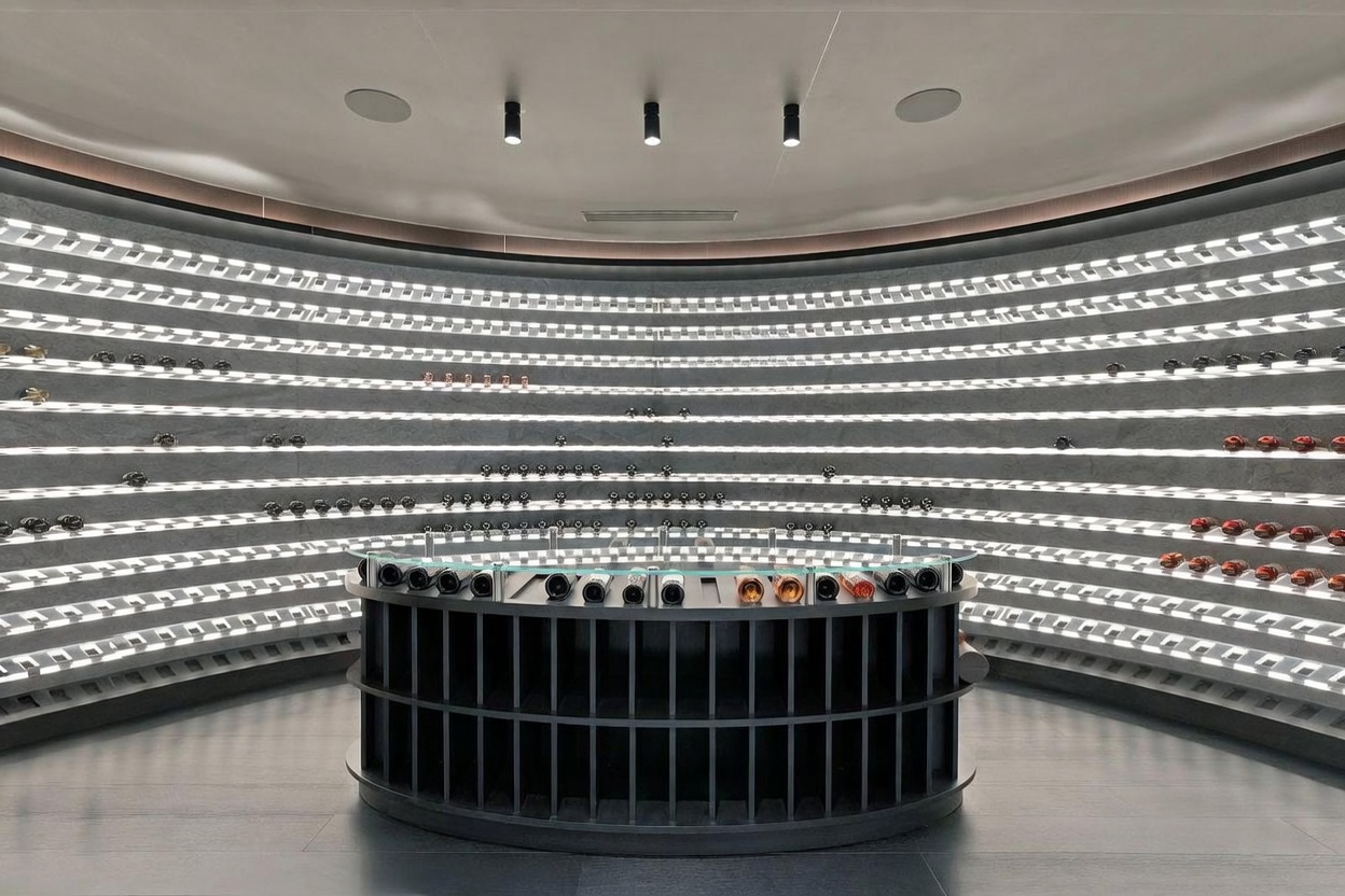 This sophisticated, circular wine cellar features a dramatic curved wall lined with illuminated shelving for extensive bottle storage. A central, semi-circular island with a glass top provides a sleek display area, while recessed ceiling lights create a focused, gallery-like atmosphere. The minimalist design and dark color palette emphasize a modern, high-end aesthetic perfect for a luxury collection.