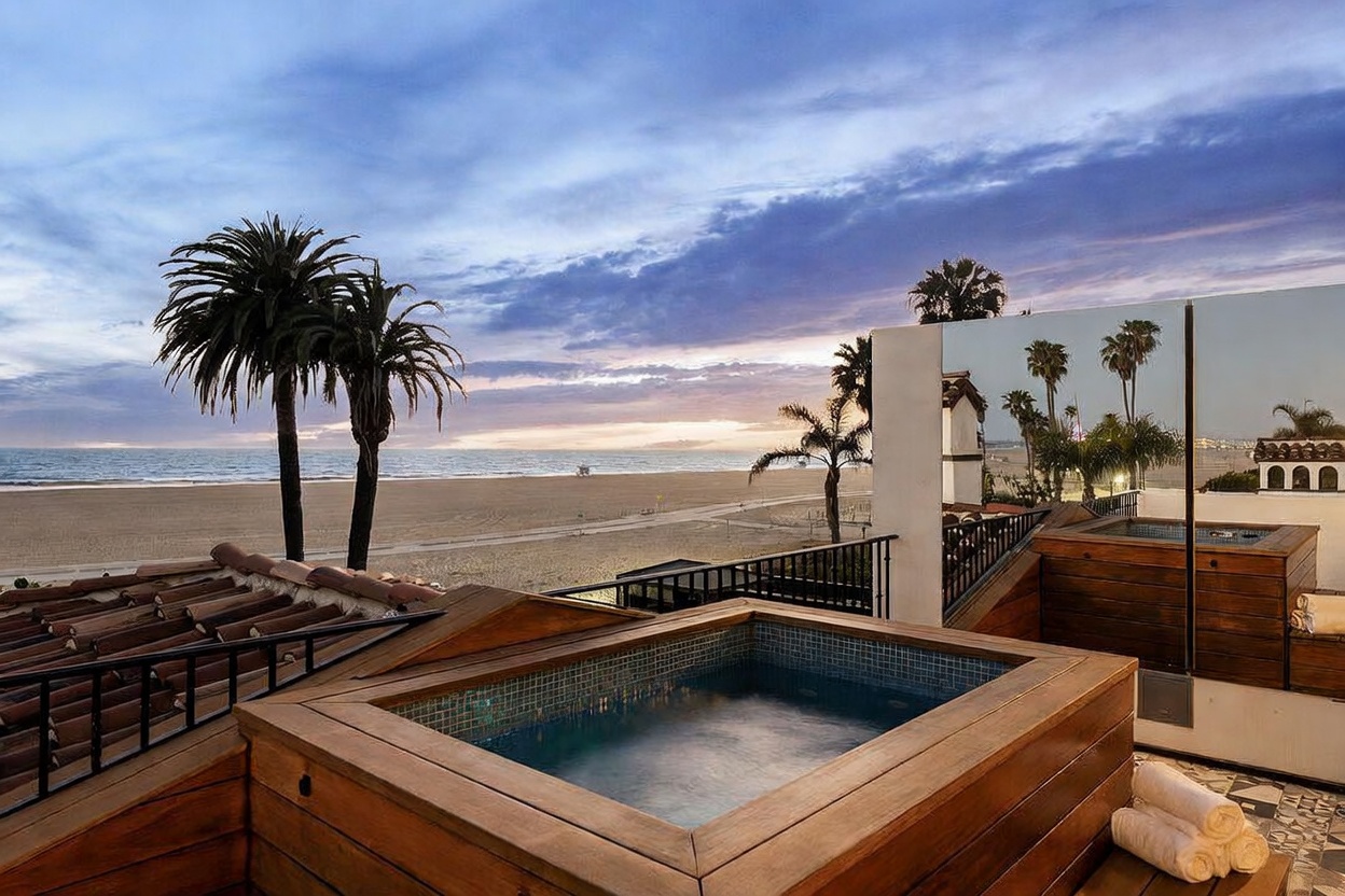 This elevated deck features a custom-built wooden spa overlooking a wide, sandy beach and the Pacific Ocean. The scene is captured at sunset, with a dramatic, cloud-filled sky providing a serene backdrop for the outdoor living space. A reflective glass partition adds a modern touch, mirroring the surrounding palm trees and coastal environment.