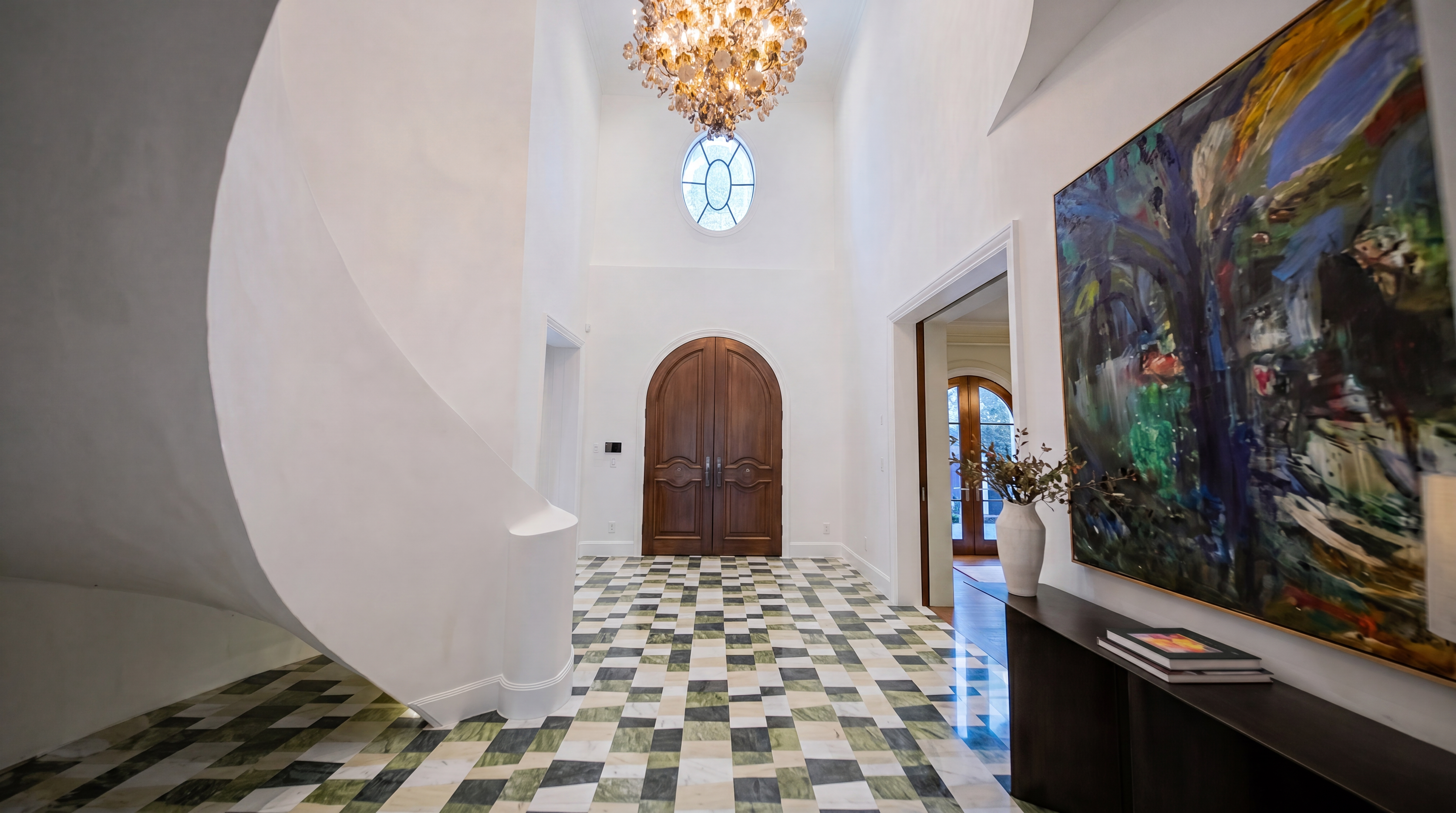 This grand foyer features a dramatic, sweeping curved staircase and a striking checkerboard-patterned marble floor. An elegant chandelier hangs from the high ceiling, complementing the arched wooden double doors and the large, abstract piece of art displayed on the wall. The space projects a luxurious and artistic atmosphere, quintessential of a high-end estate.