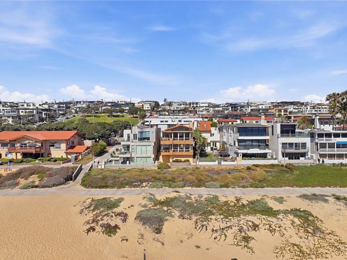 This aerial perspective captures a row of multi-story coastal residences situated directly along a sandy beach. The homes feature diverse architectural styles, including modern glass-fronted designs and traditional structures with tiled roofs, all overlooking a paved boardwalk and dunes. The bright, clear sky and expansive beach setting emphasize the prime waterfront location of these properties.