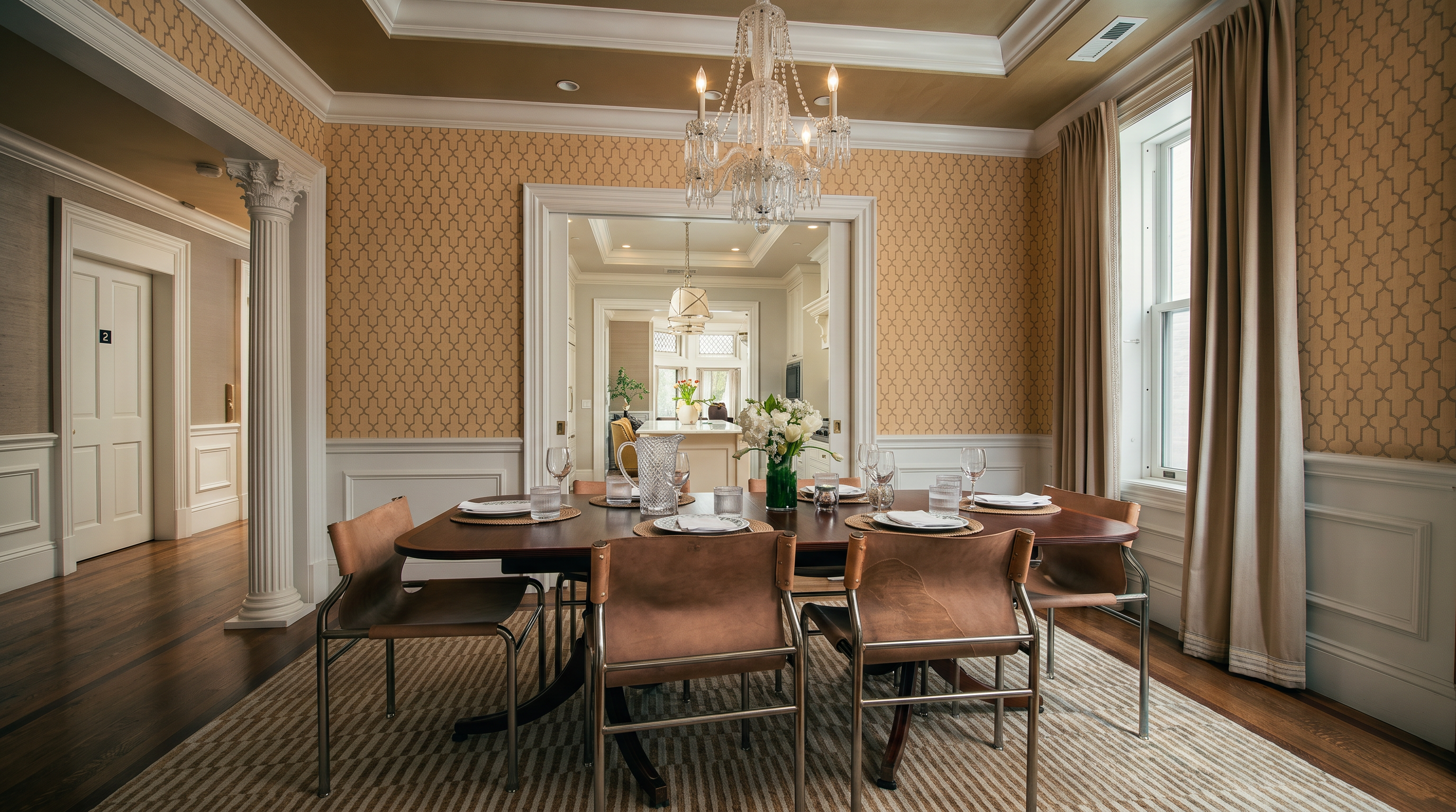 This elegant dining room features a formal wooden table surrounded by modern leather-upholstered chairs, set beneath a classic crystal chandelier. The walls are adorned with patterned wallpaper above white wainscoting, and the room is framed by a decorative column and a wide doorway leading into a bright kitchen. A large area rug anchors the space, while tall curtains frame the windows, creating a sophisticated and inviting atmosphere.