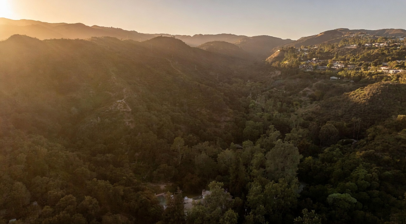 This wide-angle aerial shot captures a sprawling, lush canyon landscape bathed in the warm, golden light of sunset. The perspective highlights the dense, rolling hills covered in native vegetation, with a secluded residential property nestled deep within the valley floor. The scene emphasizes the privacy and natural beauty of the location, showcasing the expansive terrain and the serene, mountainous backdrop.